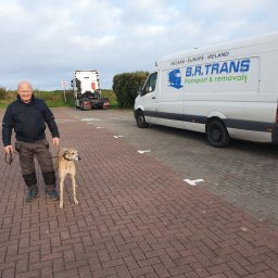 FHU B.R.Trans Ryszard Szmyglewski - Mężczyzna z psem rasy chart stoi na parkingu obok białego busa z napisem B.R. TRANS transport & removals, w tle widoczna ciężarówka.