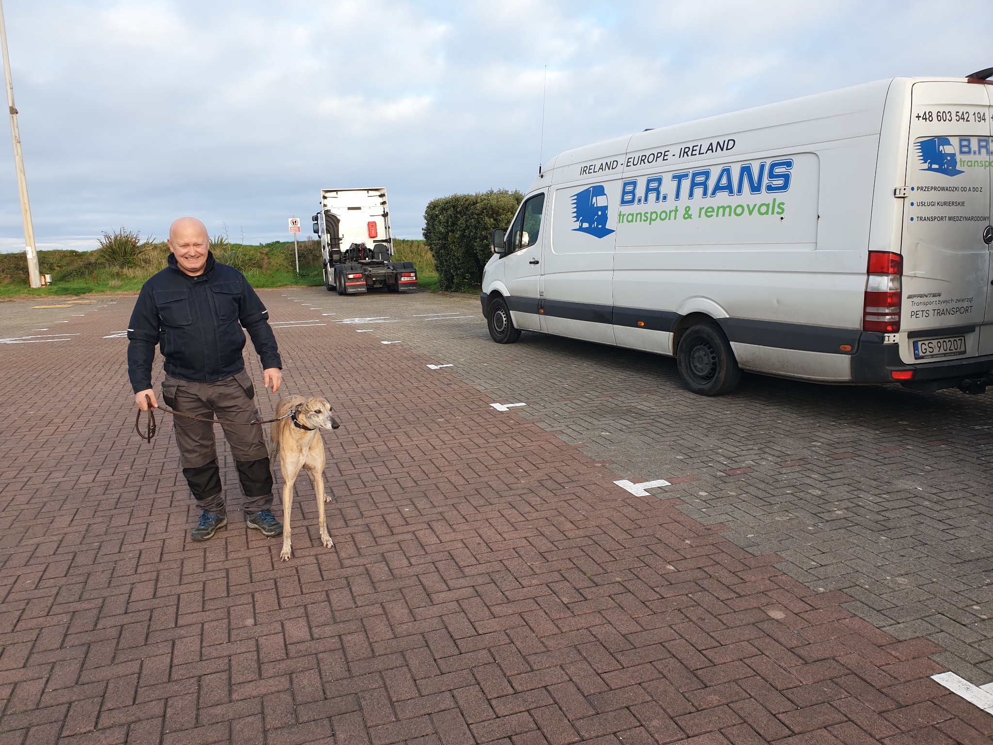 Mężczyzna z psem rasy chart stoi na parkingu obok białego busa z napisem B.R. TRANS transport & removals, w tle widoczna ciężarówka.