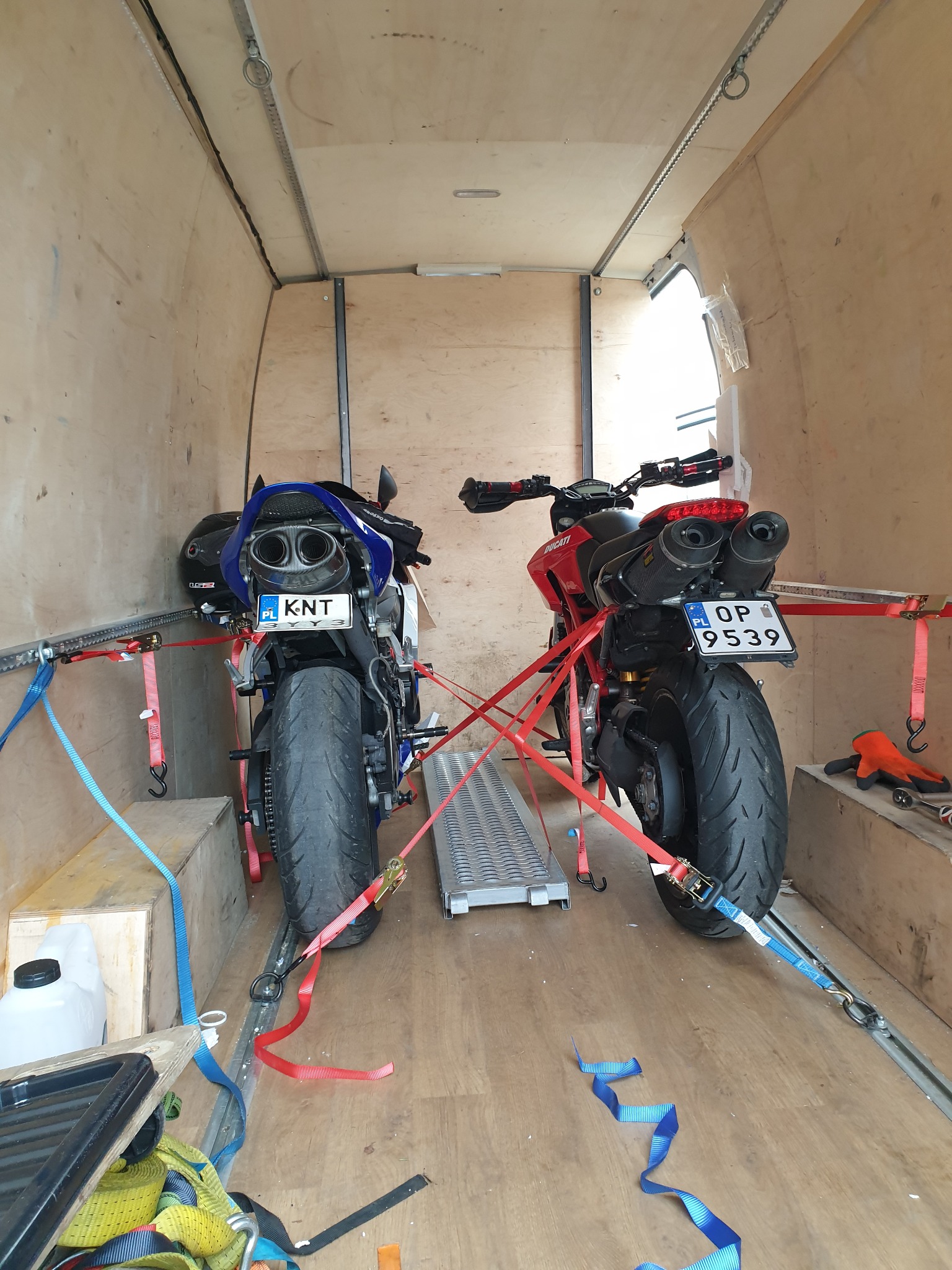 Dwa motocykle, czerwony Ducati i niebieski Yamaha, zabezpieczone pasami wewnątrz drewnianej furgonetki, gotowe do transportu. Widoczne tablice rejestracyjne i rampy najazdowe.