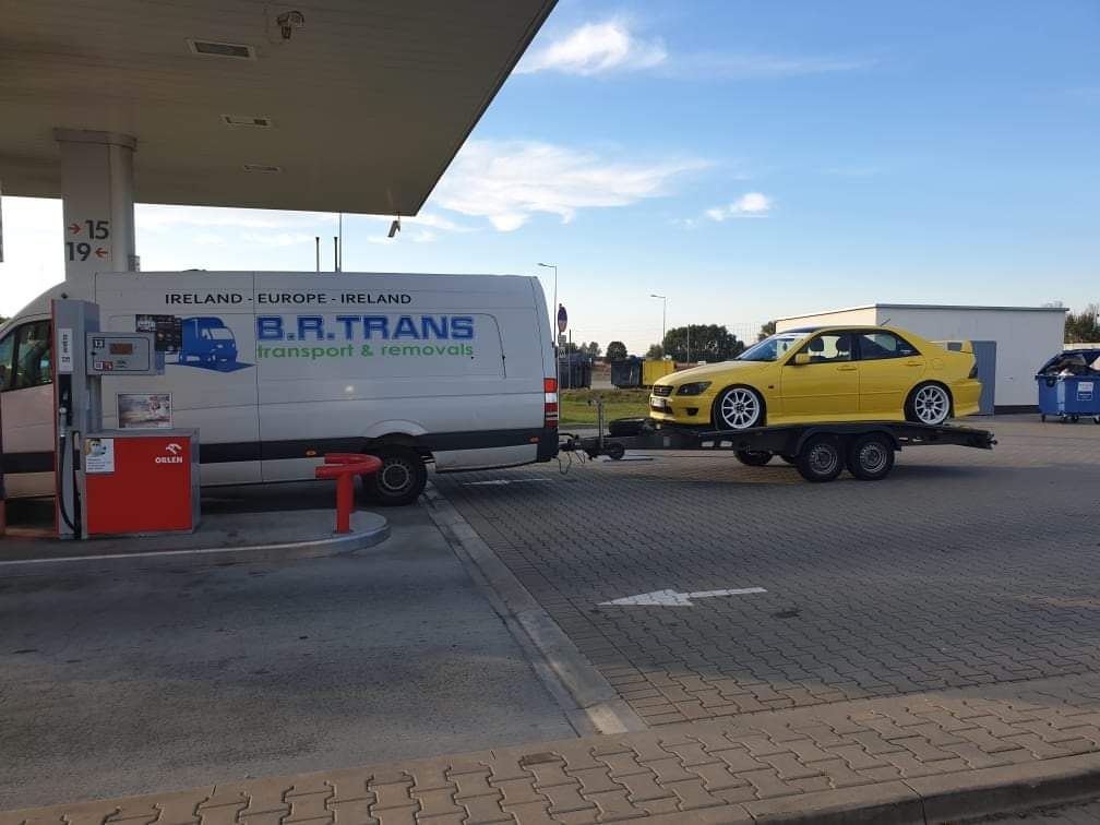 Żółty samochód sportowy na lawecie, ciągnięty przez biały bus firmy B.R. Trans na stacji benzynowej Orlen.