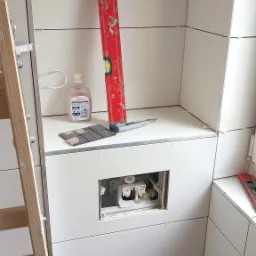 Białe płytki układane w łazience, widoczny poziomica, nożyk, mydło w płynie oraz otwór na stelaż podtynkowy WC.