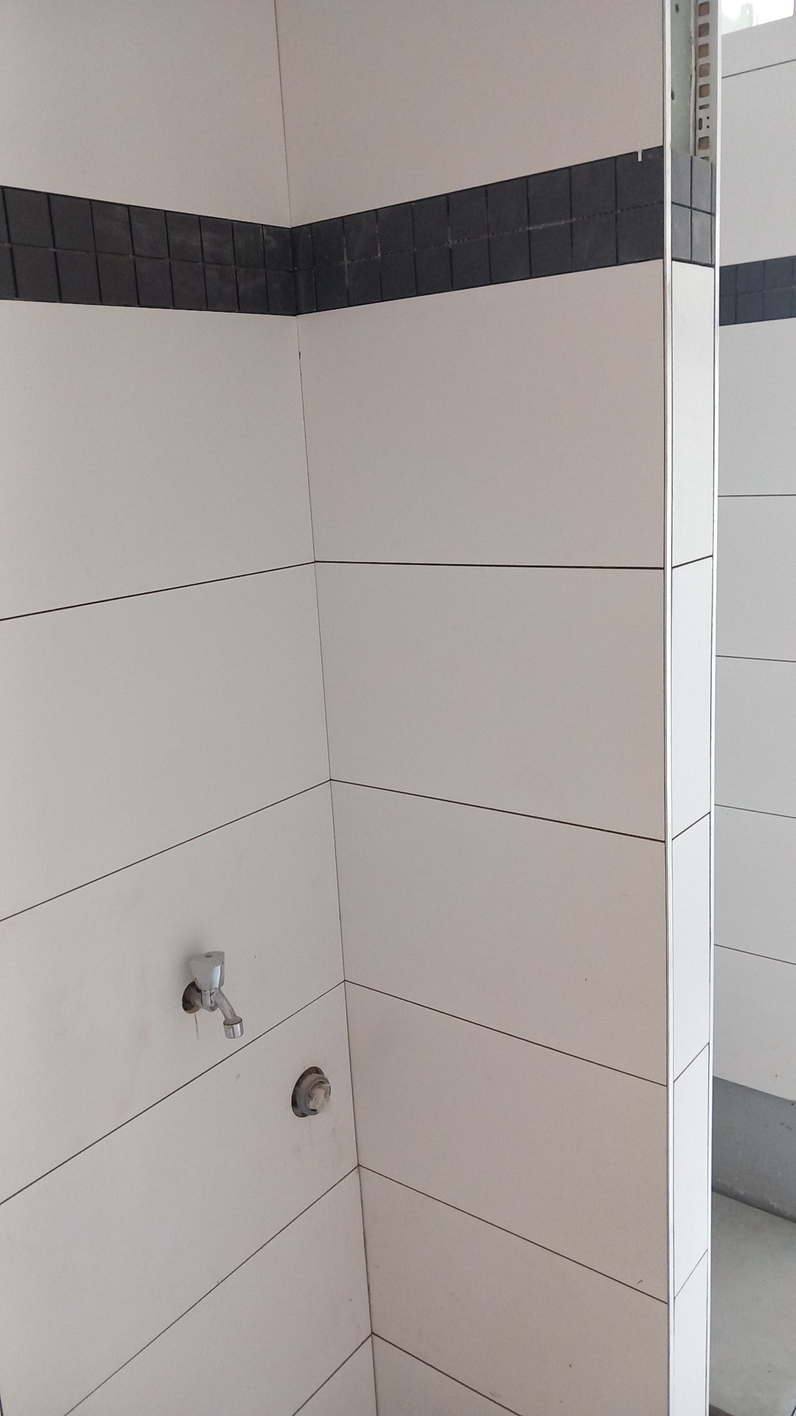 Ściana łazienki wyłożona białymi, poziomymi płytkami z czarnym pasem mozaiki u góry, widoczne przygotowane otwory na armaturę.