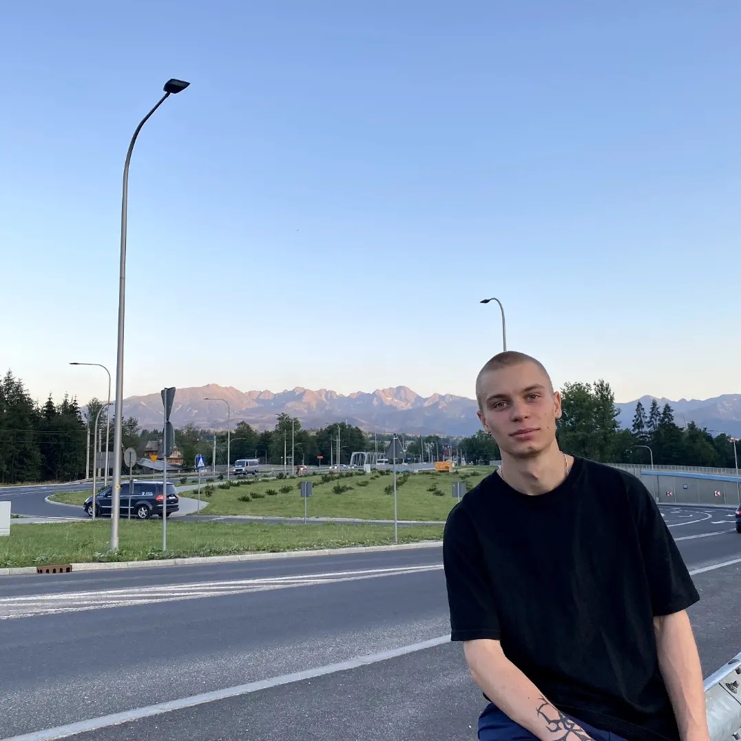 Młody mężczyzna z tatuażem na ramieniu siedzi na barierce przy drodze z widokiem na góry w oddali, oświetlony popołudniowym słońcem.