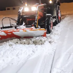 Ciężki traktor z pomarańczowym pługiem odśnieżającym usuwa śnieg z drogi nocą, widoczne ślady opon w śniegu.
