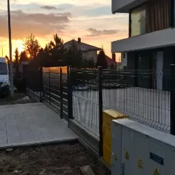 Nowoczesne ogrodzenie panelowe z bramą, widok z zewnątrz, szara kostka brukowa, skrzynki gazowe, dom w tle, zachód słońca, ciepłe światło.