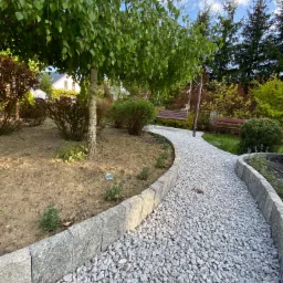 Kamyczkowa ścieżka w ogrodzie z obrzeżami z szarej kostki granitowej, prowadząca do huśtawki i ławki pod drzewami, na tle zieleni i błękitnego nieba.