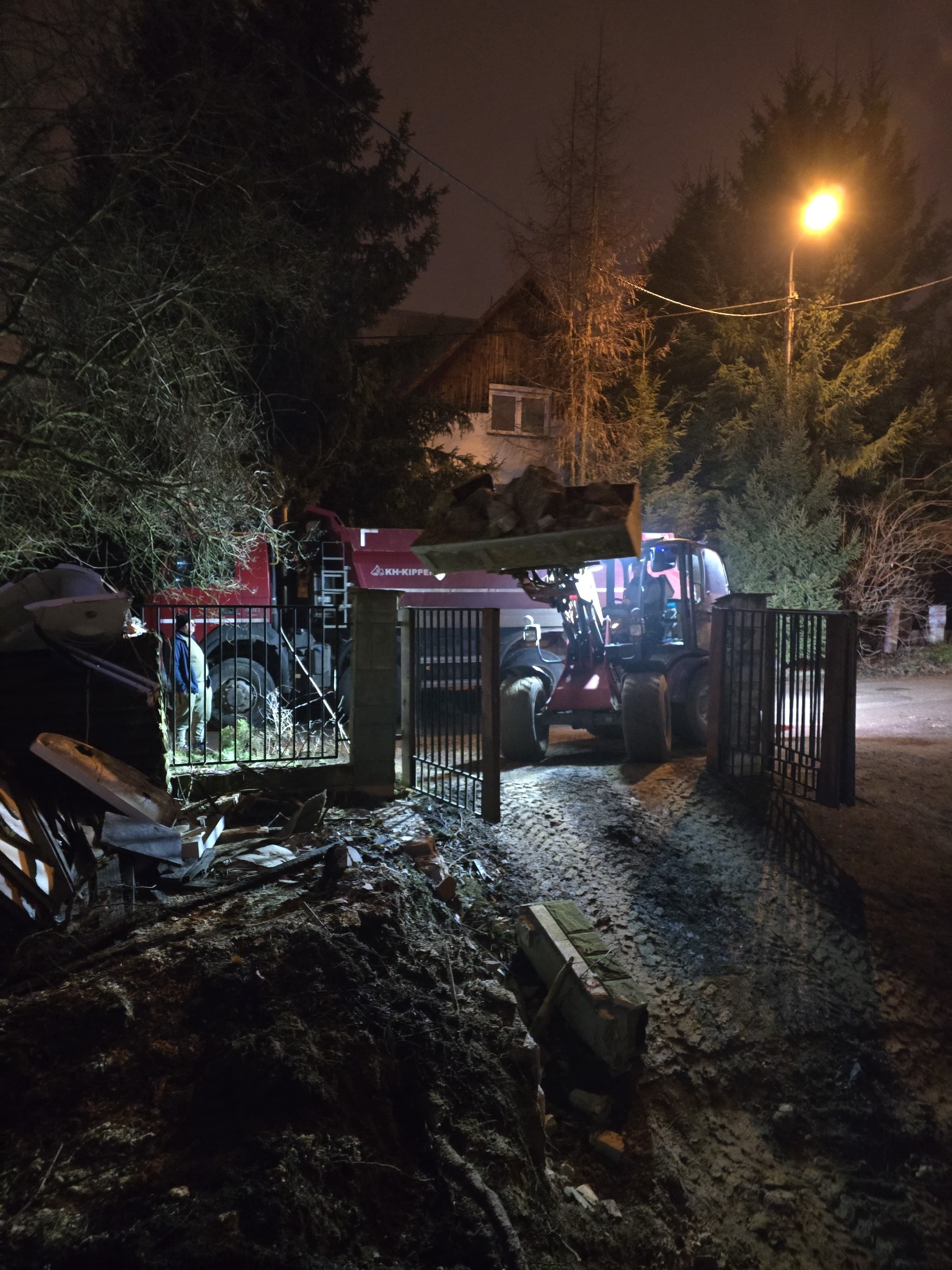 Czerwony teleskopowy ładowacz przez otwartą bramę wjeżdża na teren budowy, w tle widoczne drzewa i oświetlona latarnią ulica, po lewej sterta gruzu.