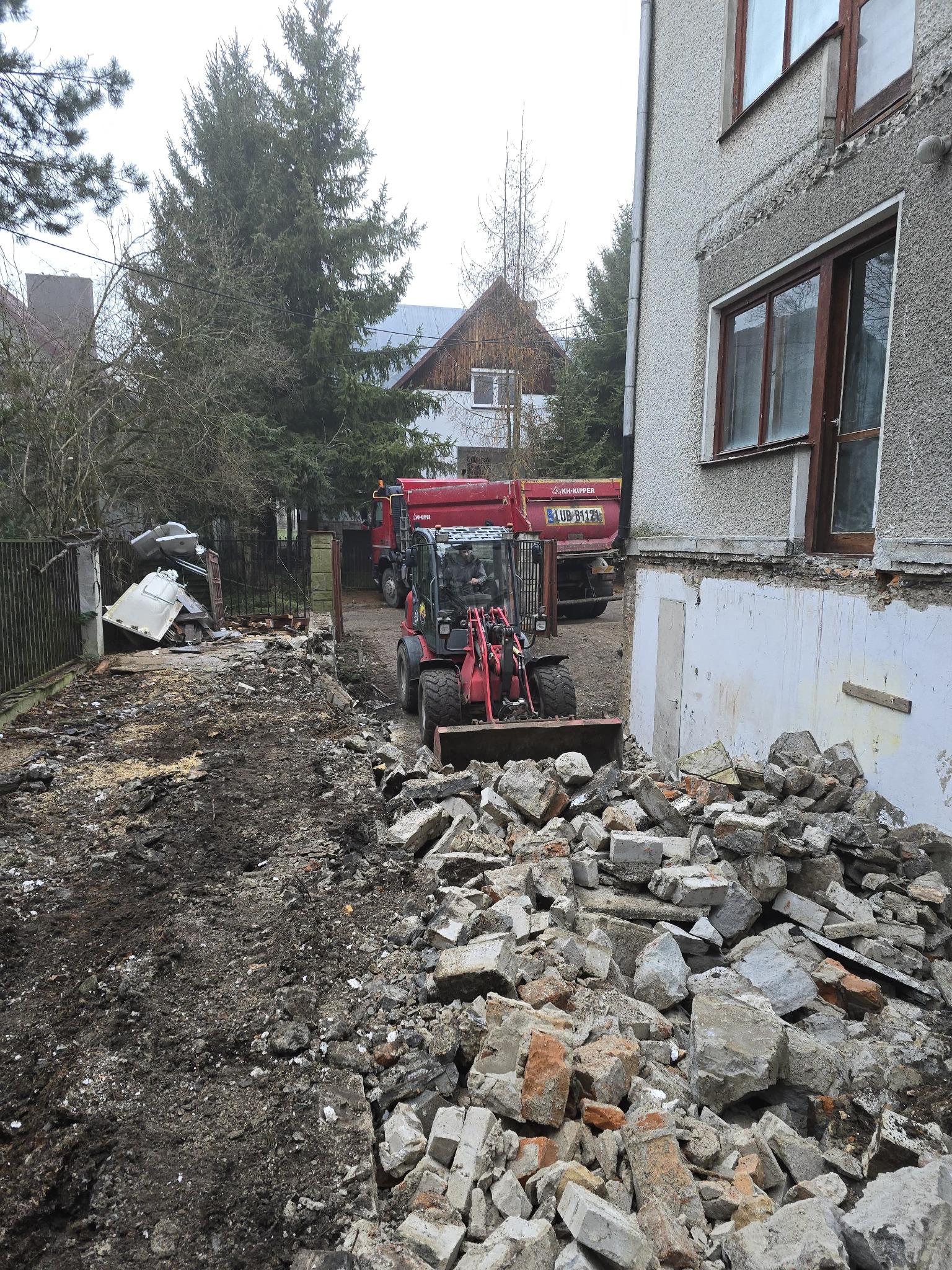 Miniładowarka z łyżką pełną gruzu przed budynkiem w trakcie prac rozbiórkowych, widoczna ciężarówka i resztki zniszczonego ogrodzenia.