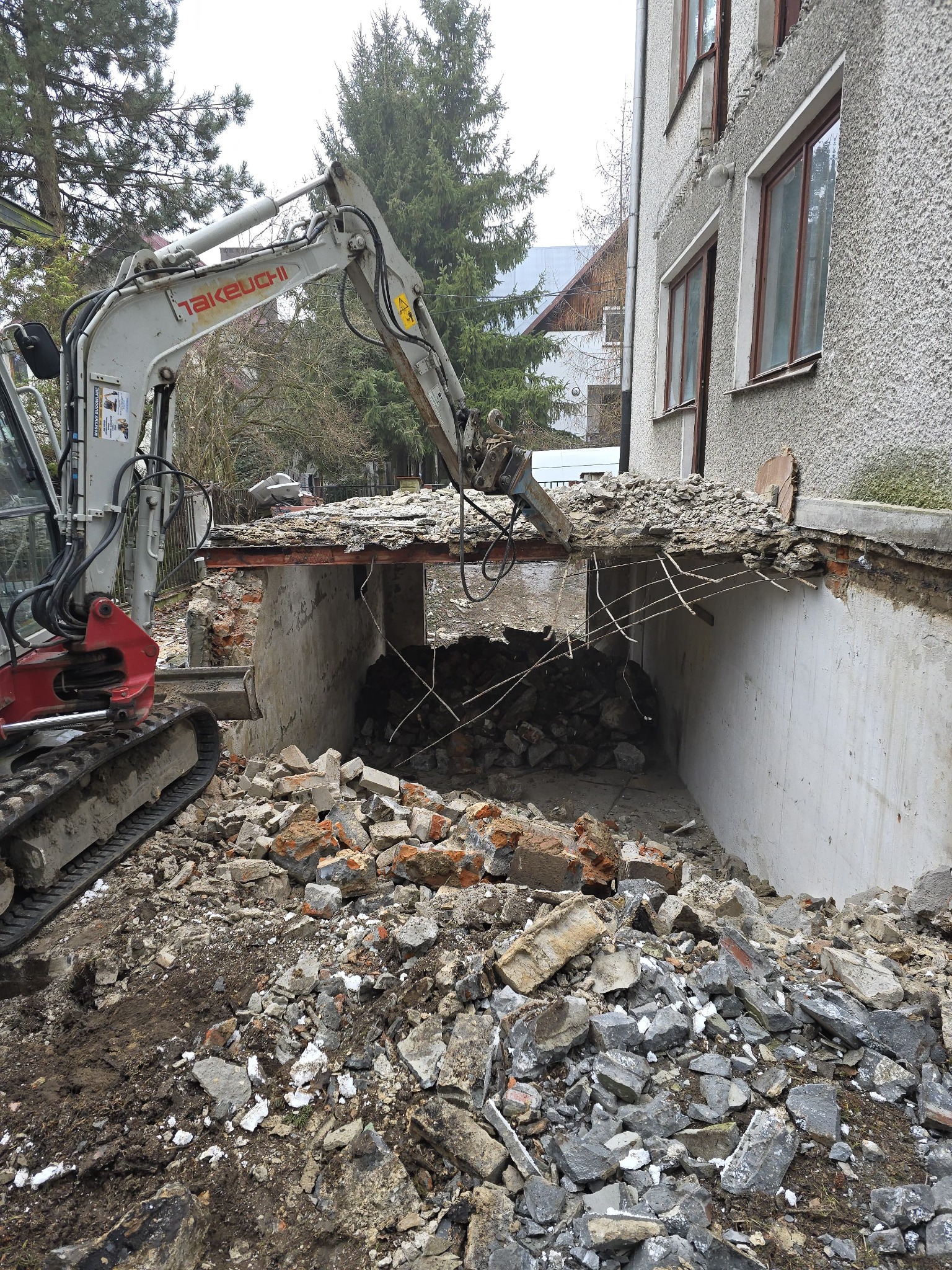 Mini koparka Takeuchi z hydraulicznym młotem kruszy betonową konstrukcję budynku, gruz i ziemia na pierwszym planie.