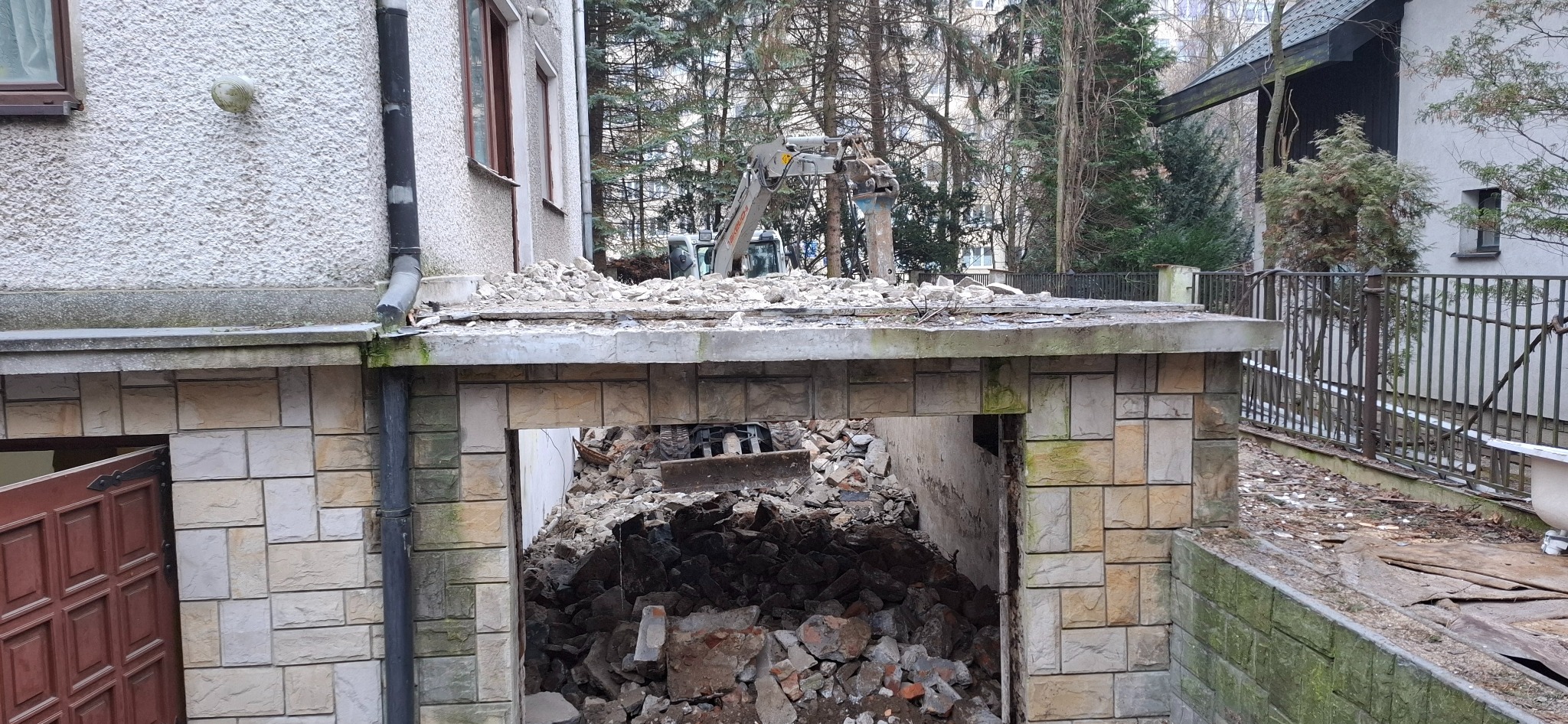 Częściowo zburzony budynek z gruzem, widoczna koparka z przodu budynku i resztki materiałów budowlanych.