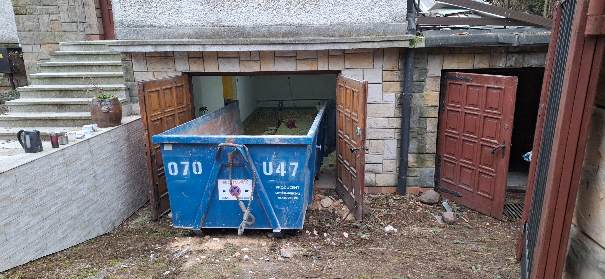 Niebieski kontener na gruz z numerami 070 i U47, stojący przed otwartym garażem z widocznymi resztkami gruzu w środku, obok uchylone drewniane drzwi, fragment schodów i donica z rośliną.