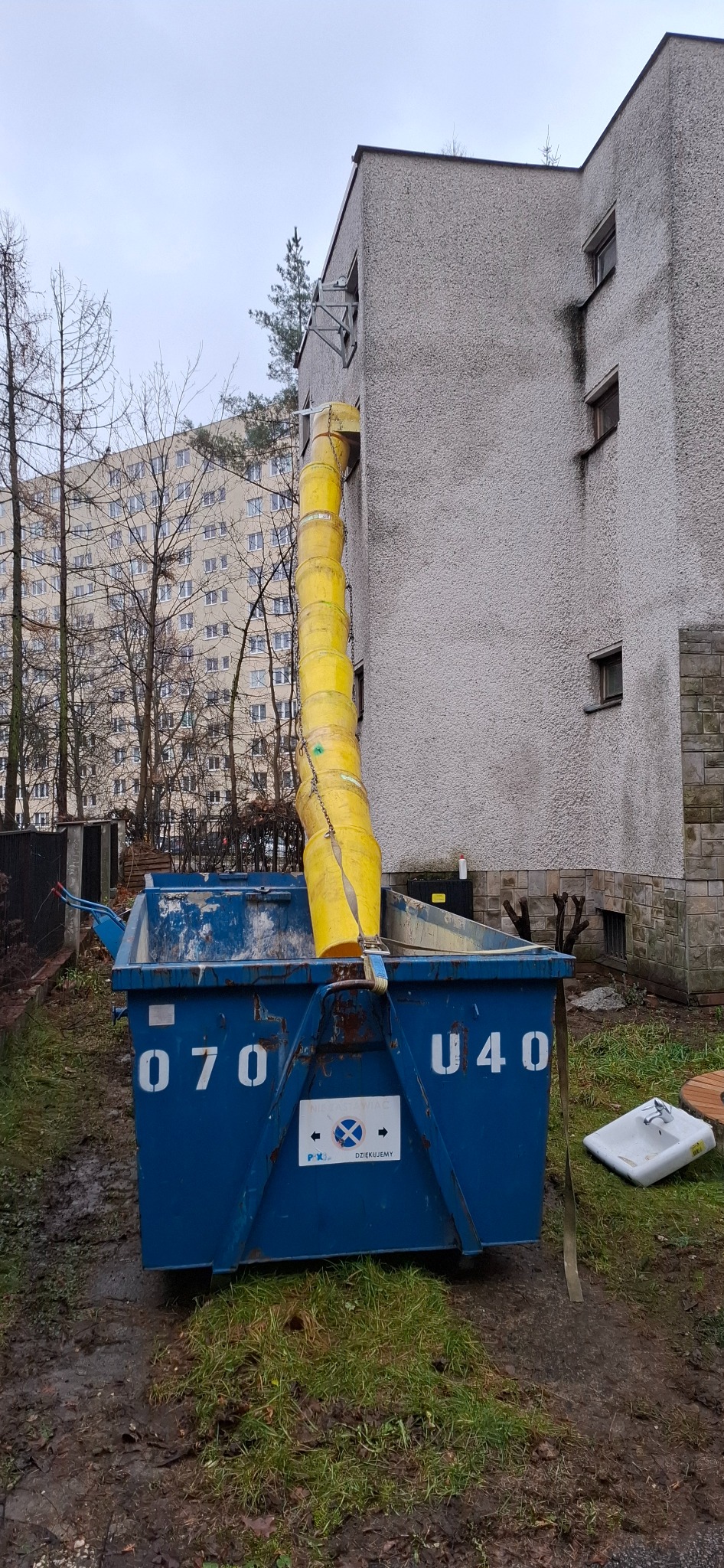 Żółty rękaw zsypowy przymocowany do budynku, prowadzący do niebieskiego kontenera na gruz. Na ziemi widoczne resztki gruzu i stara umywalka.