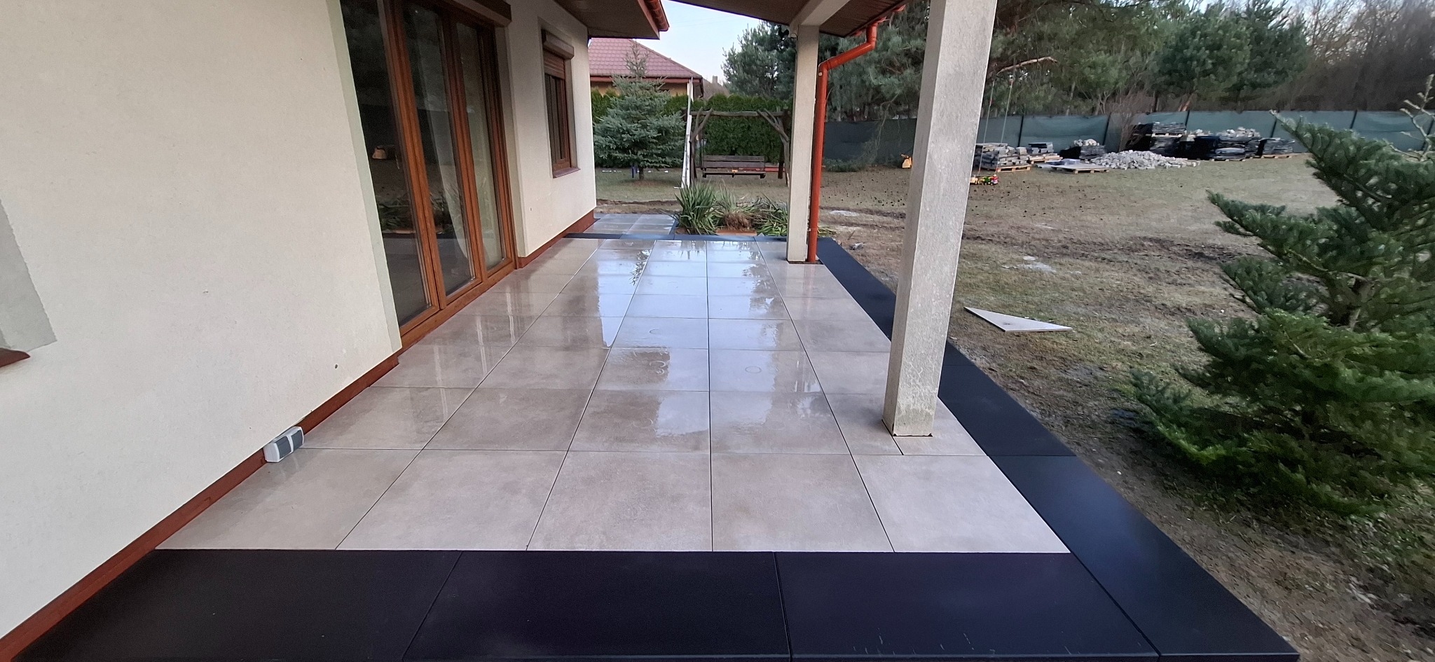 Wyłożony jasnymi płytami taras z czarnym obramowaniem, widok z perspektywy, w tle huśtawka ogrodowa i palety z materiałami budowlanymi.