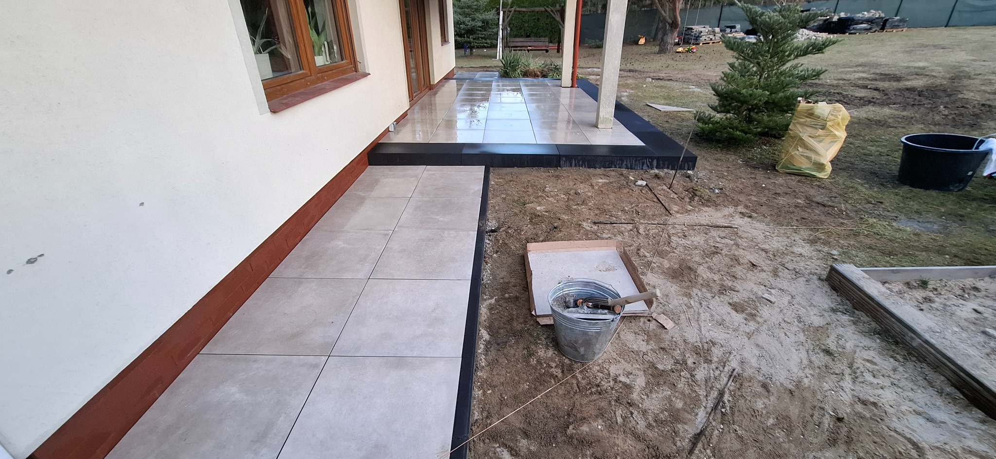 Jasnoszare płytki ułożone wzdłuż ściany domu, z czarnym obramowaniem, widoczny kubeł z zaprawą i narzędzia murarskie na ziemi, w tle ogród z drzewami.