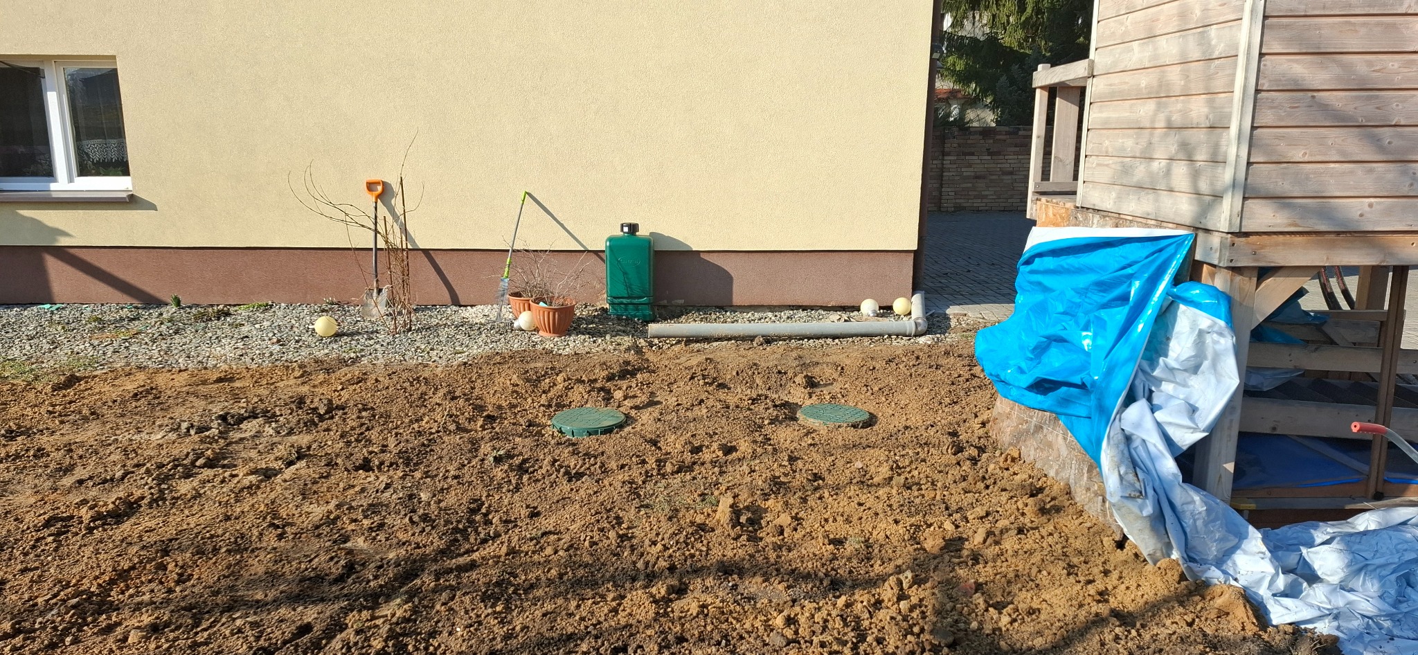 Przygotowany teren pod przyszły ogród z widocznymi dwoma włazami do instalacji podziemnej, narzędzia ogrodnicze opierają się o ścianę budynku, obok drewniana konstrukcja zabezpieczona plandeką.