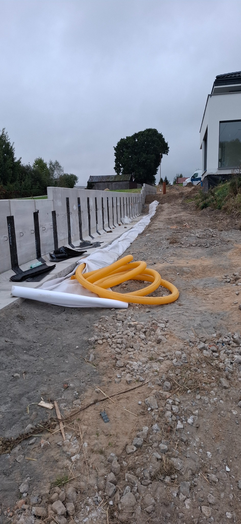 Betonowe elementy oporowe z mocowaniami, żółta rura drenarska i biała geowłóknina na placu budowy. Widok na teren z ziemią i kamieniami.