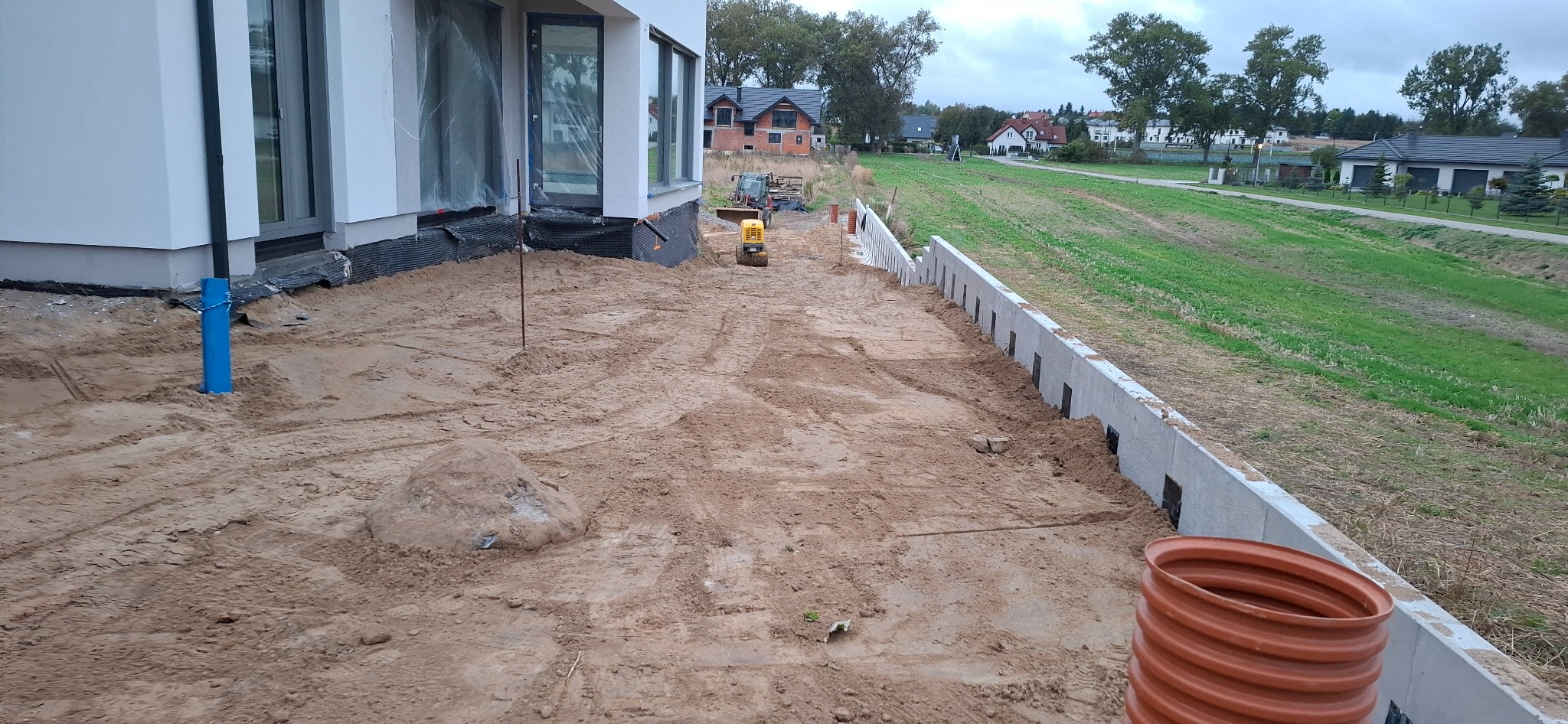 Przygotowanie terenu pod budowę: wyrównana ziemia, betonowy mur oporowy, widoczna zagęszczarka, rura kanalizacyjna. Etap prac ziemnych na działce.