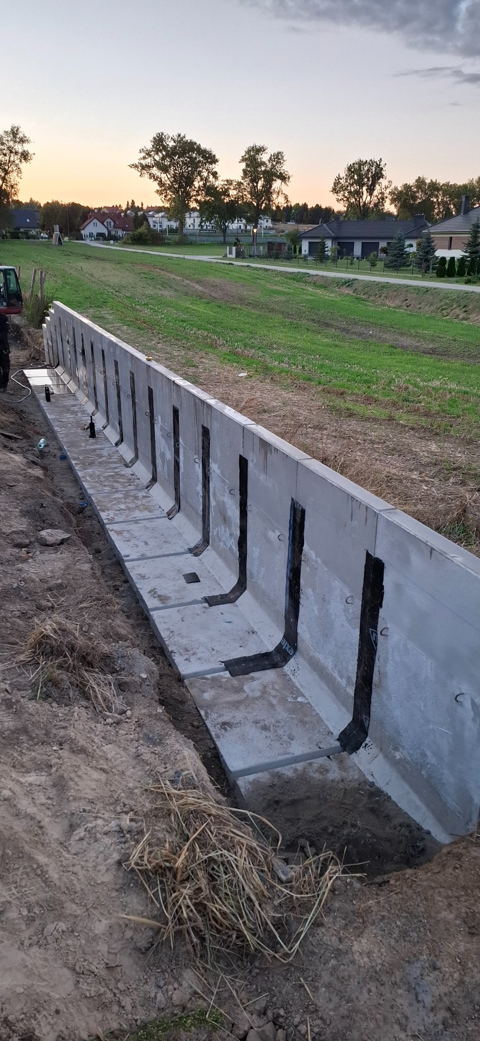 Betonowy kanał odwadniający z czarną izolacją, wkopany w ziemię, z widocznym krajobrazem i domami w tle, uchwycony podczas zachodu słońca.