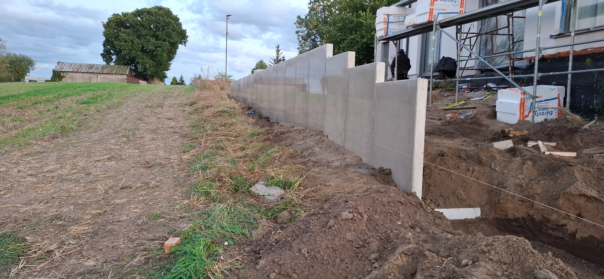 Montaż betonowego muru oporowego z charakterystycznymi uskokami, widoczne wykopy i materiały budowlane przy nowoczesnym domu w tle.