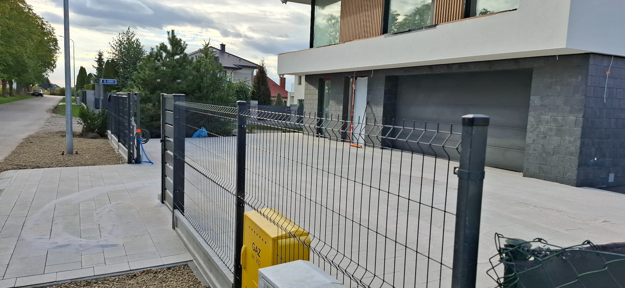 Nowoczesne ogrodzenie panelowe posesji w Lublinie, z widoczną skrzynką gazową. Szara kostka brukowa na podjeździe, nowoczesna architektura domu w tle.