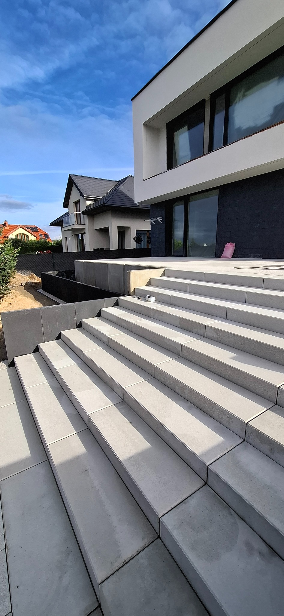 Nowoczesne schody z jasnoszarej kostki brukowej prowadzące do domu. Widoczny fragment ogrodu i sąsiedni dom. Słoneczny dzień, błękitne niebo.