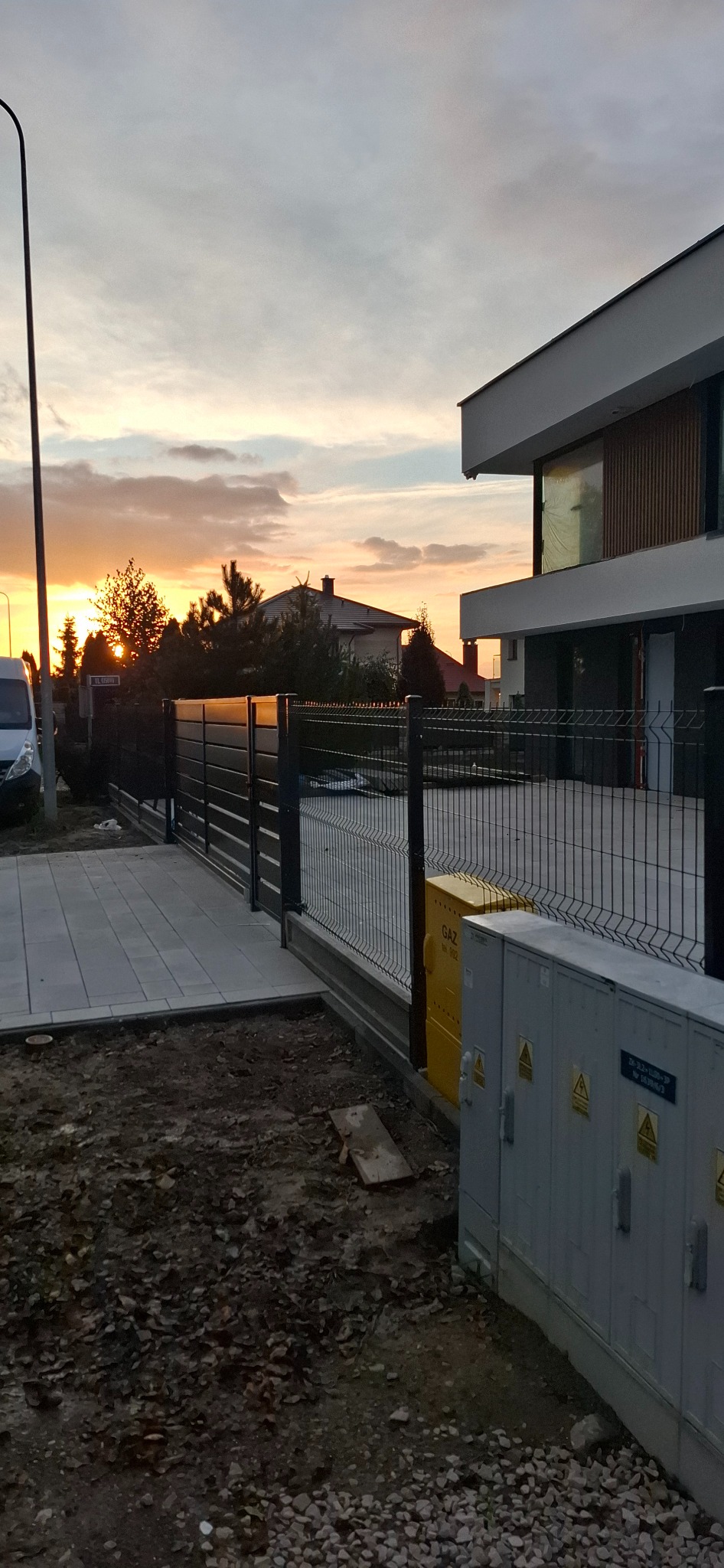 Nowoczesne ogrodzenie panelowe z bramą, widok z zewnątrz, szara kostka brukowa, skrzynki gazowe, dom w tle, zachód słońca, ciepłe światło.