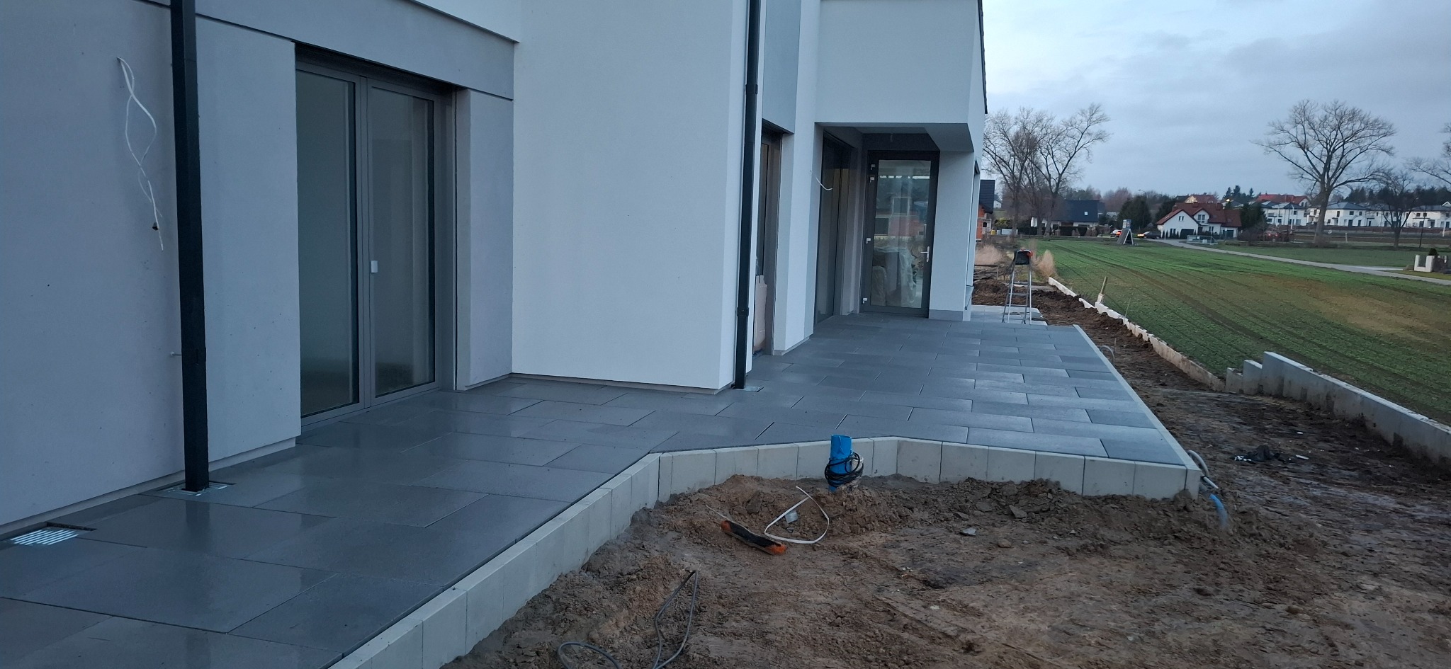 Nowoczesny taras z szarej kostki brukowej przy białym domu. Widoczne narzędzia i przygotowany teren pod ogród. Minimalistyczny design i stonowana kolorystyka.