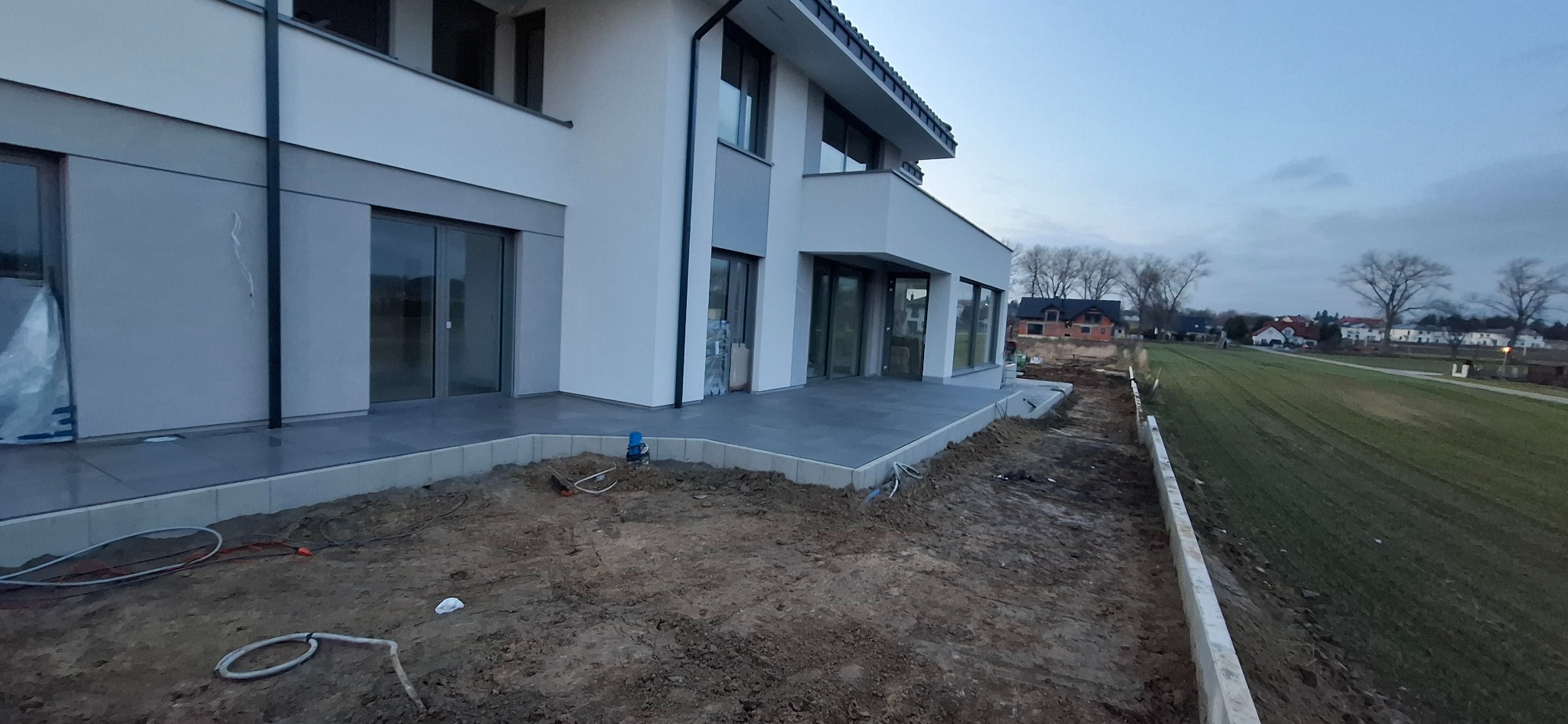 Nowoczesny taras z szarych płyt przy białym domu jednorodzinnym. Widoczne przygotowania do dalszych prac ziemnych i wykończeniowych wokół tarasu.