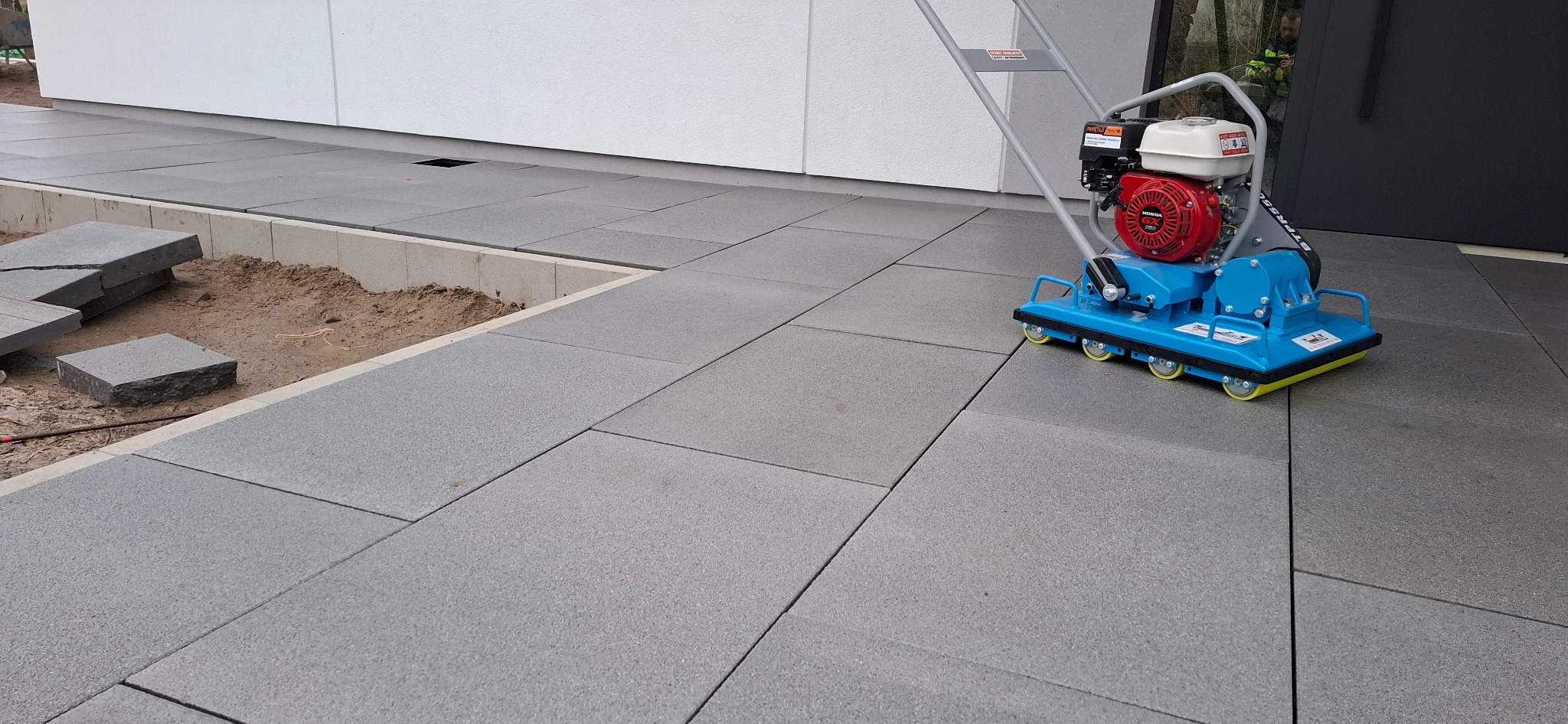 Świeżo ułożona, szara kostka brukowa tworzy ścieżkę. W tle biała ściana budynku i niebieska zagęszczarka wibracyjna. Część kostki jeszcze nieułożona.