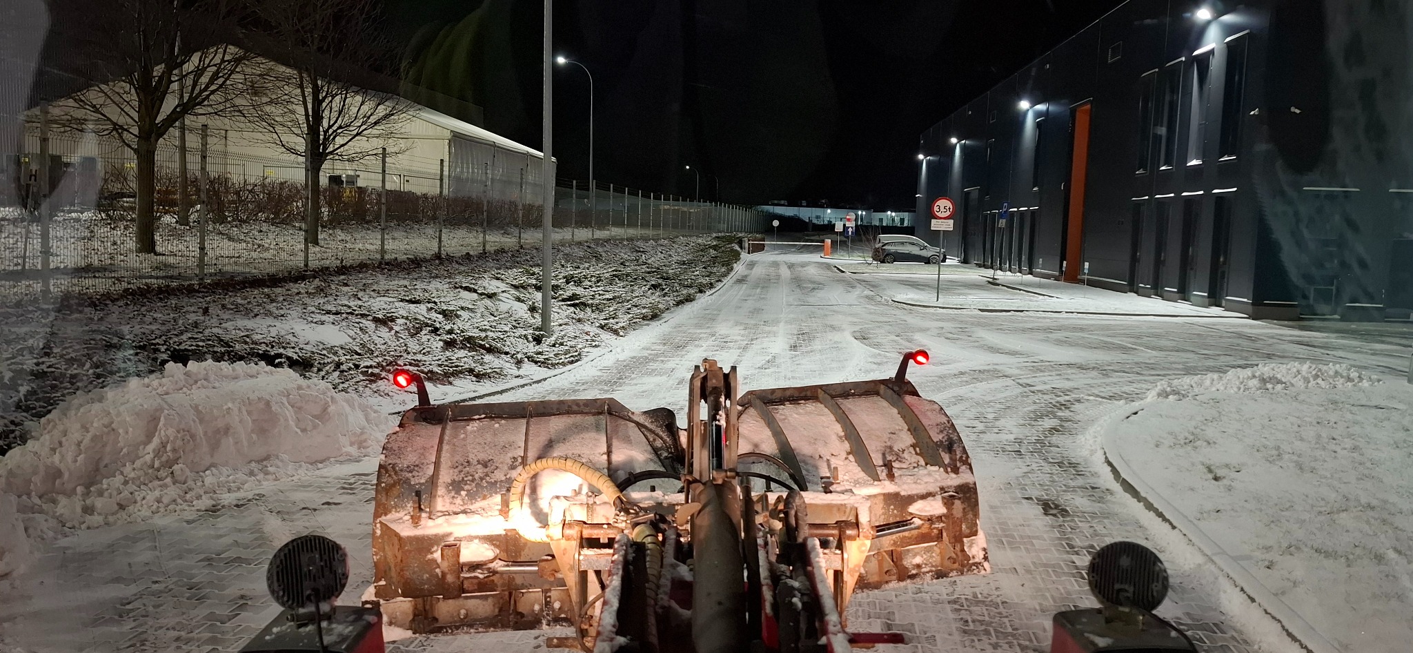Odśnieżarka w akcji, usuwająca śnieg z drogi w nocy. Widoczne budynki w tle i oświetlenie uliczne. Świeży śnieg zalega na powierzchni.