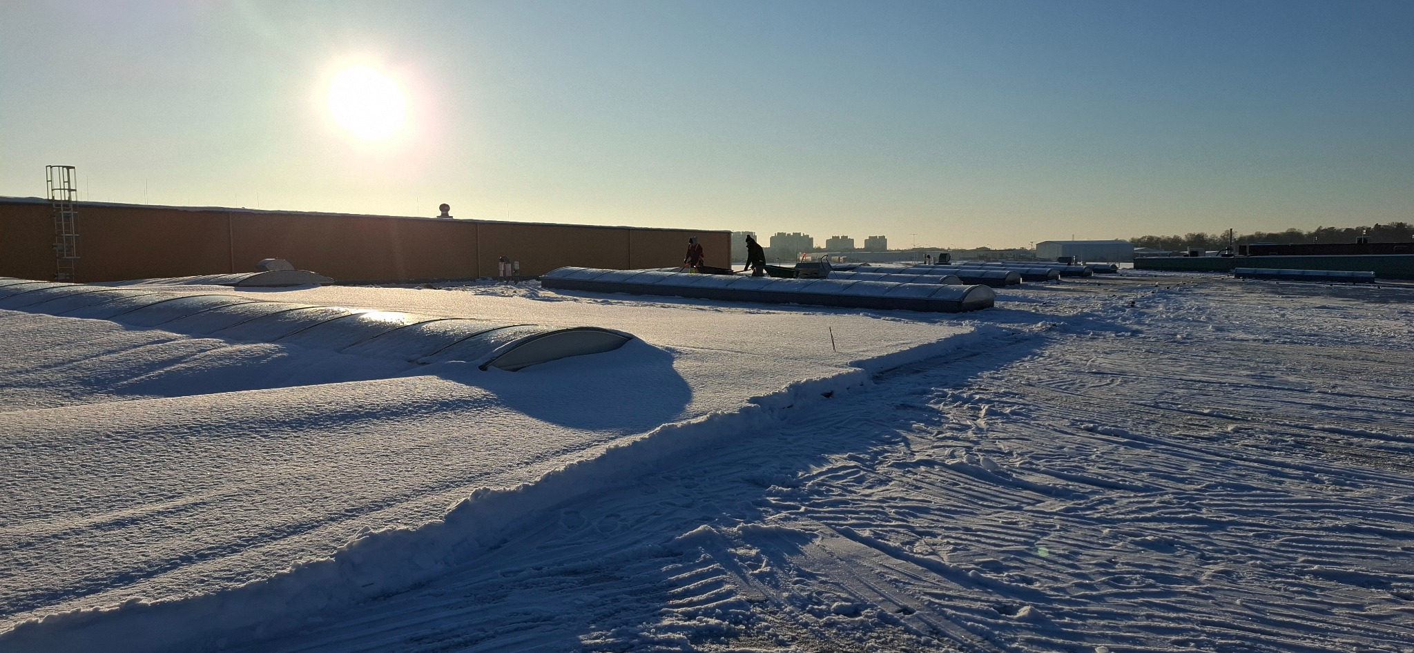 Odśnieżanie dachu budynku z świetlikami. Widoczne ślady po odśnieżaniu i pracownicy przy pracy. Słoneczny dzień.