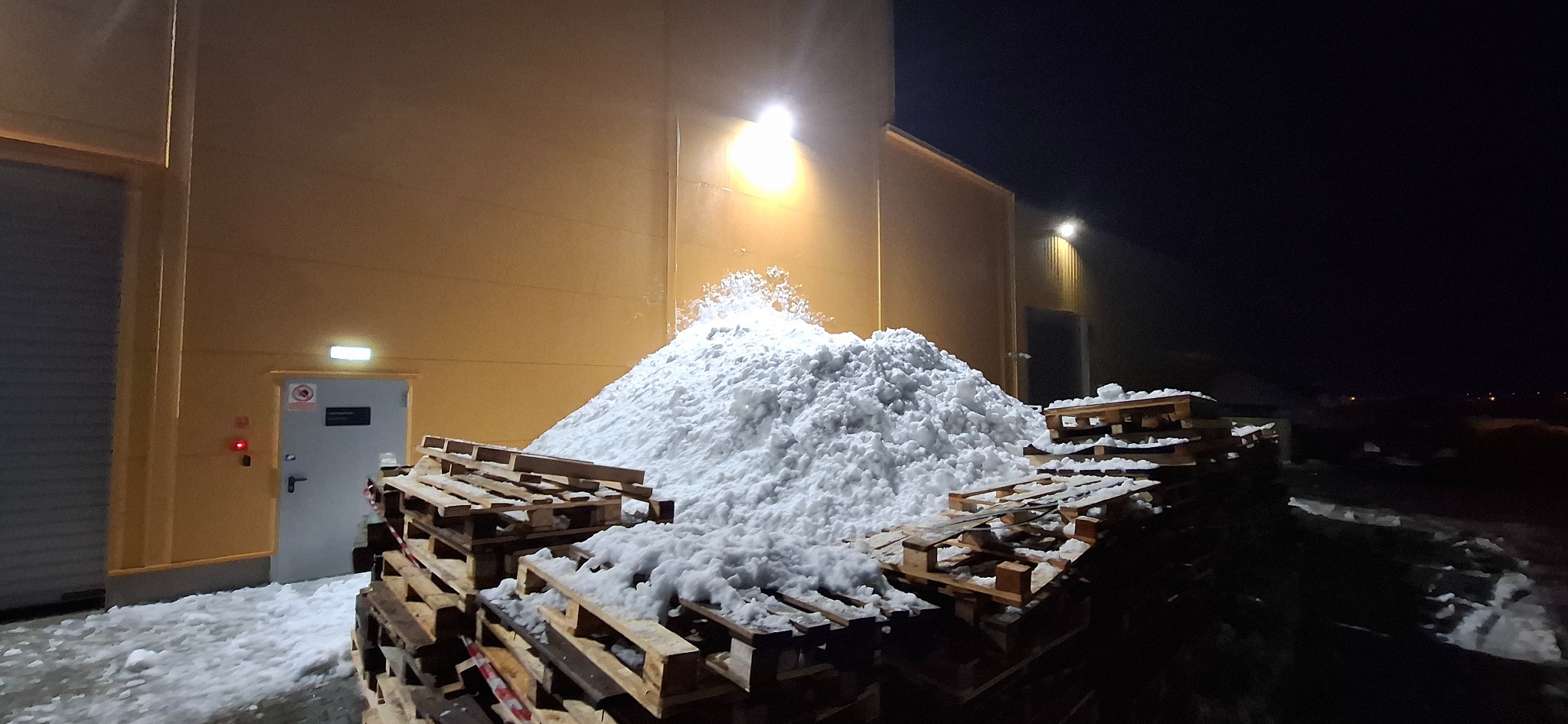 Góra śniegu na paletach przy budynku w Lublinie, oświetlona nocą. Widoczne drzwi wejściowe i lampy na elewacji. Śnieg sprawia wrażenie świeżo odgarniętego.