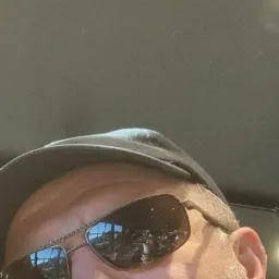 Portret mężczyzny w czapce i okularach przeciwsłonecznych, w których odbija się wnętrze pomieszczenia z oknami i stołami.