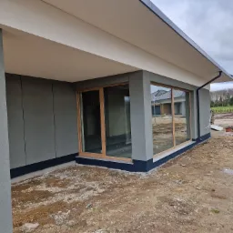 Szara elewacja domu jednorodzinnego z dużymi oknami w drewnianych ramach, widok z zewnątrz, teren wokół domu w trakcie prac wykończeniowych.