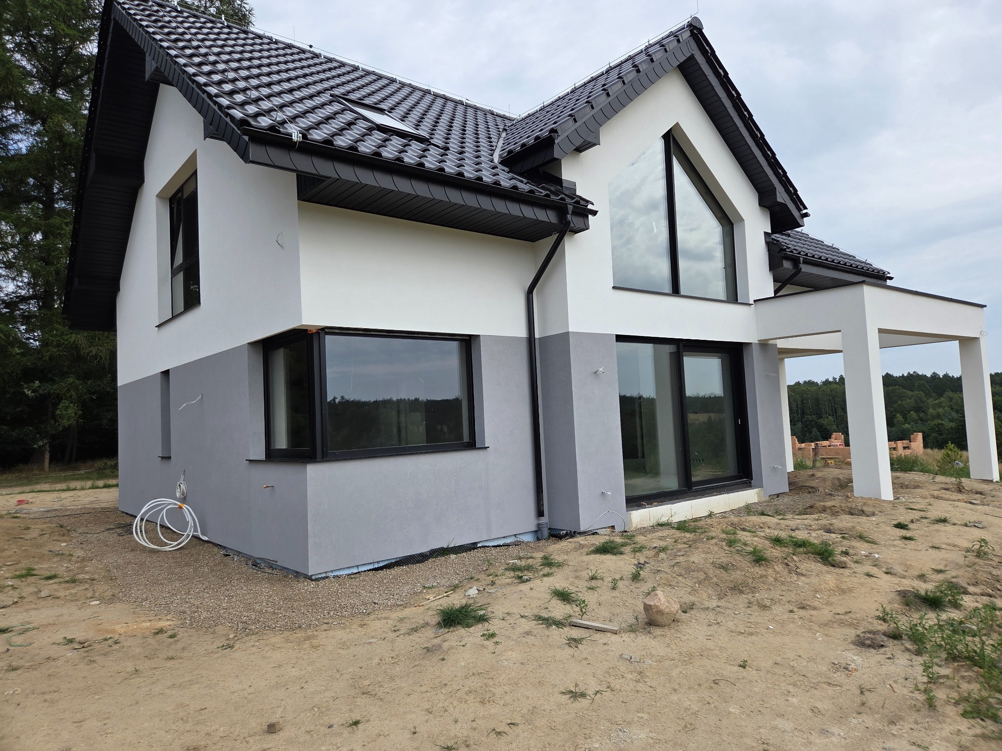 Nowoczesny dom z dwukolorową elewacją (biało-szarą), czarną dachówką i dużymi oknami. Widok z zewnątrz na plac budowy, z widocznymi elementami wykończeniowymi.
