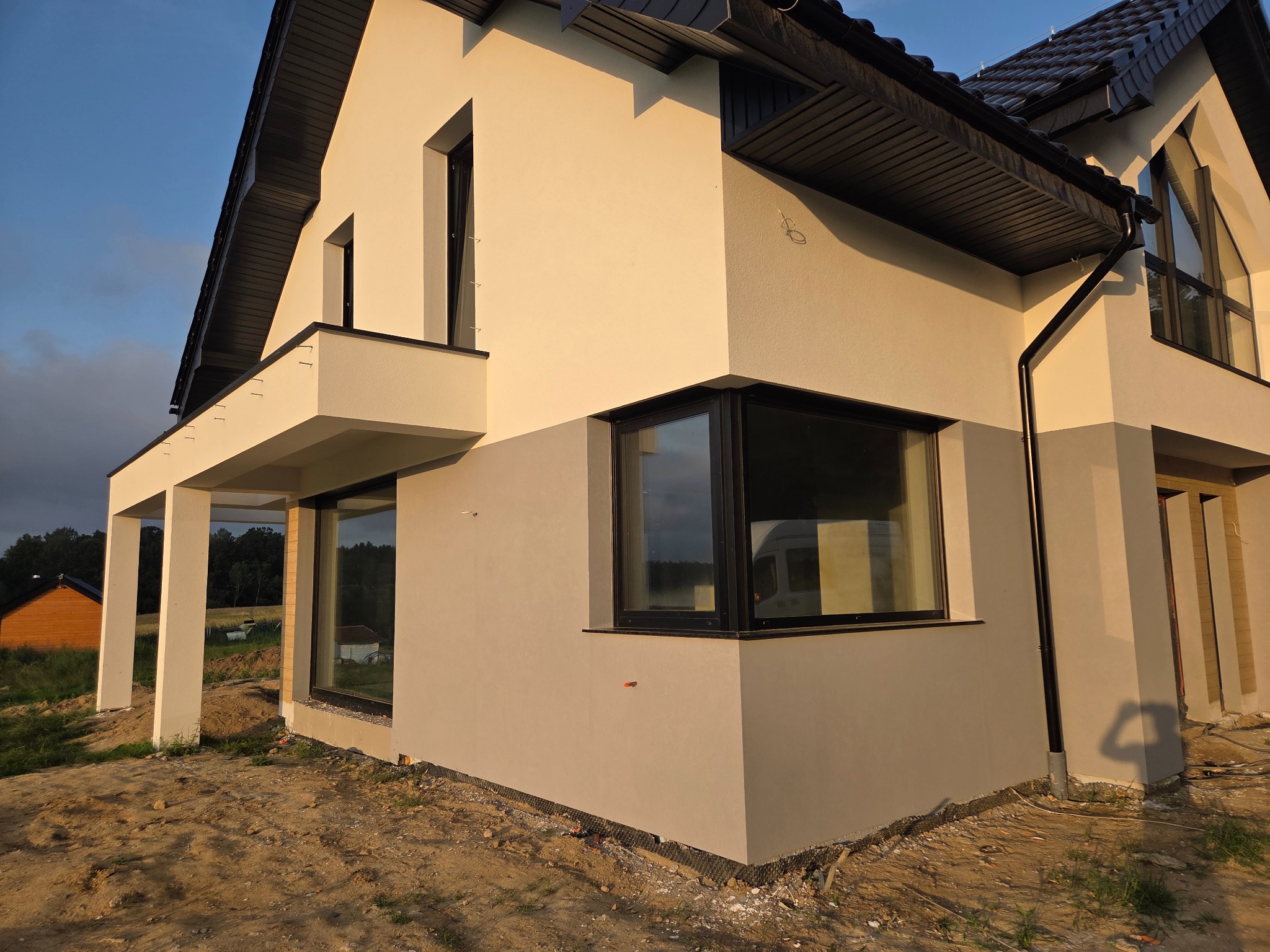 Dwukondygnacyjny dom z jasną elewacją i czarnymi oknami, widoczny z bliska z boku, w otoczeniu ziemi i trawy. Część elewacji w odcieniu szarości. Widoczne elementy wykończeniowe.