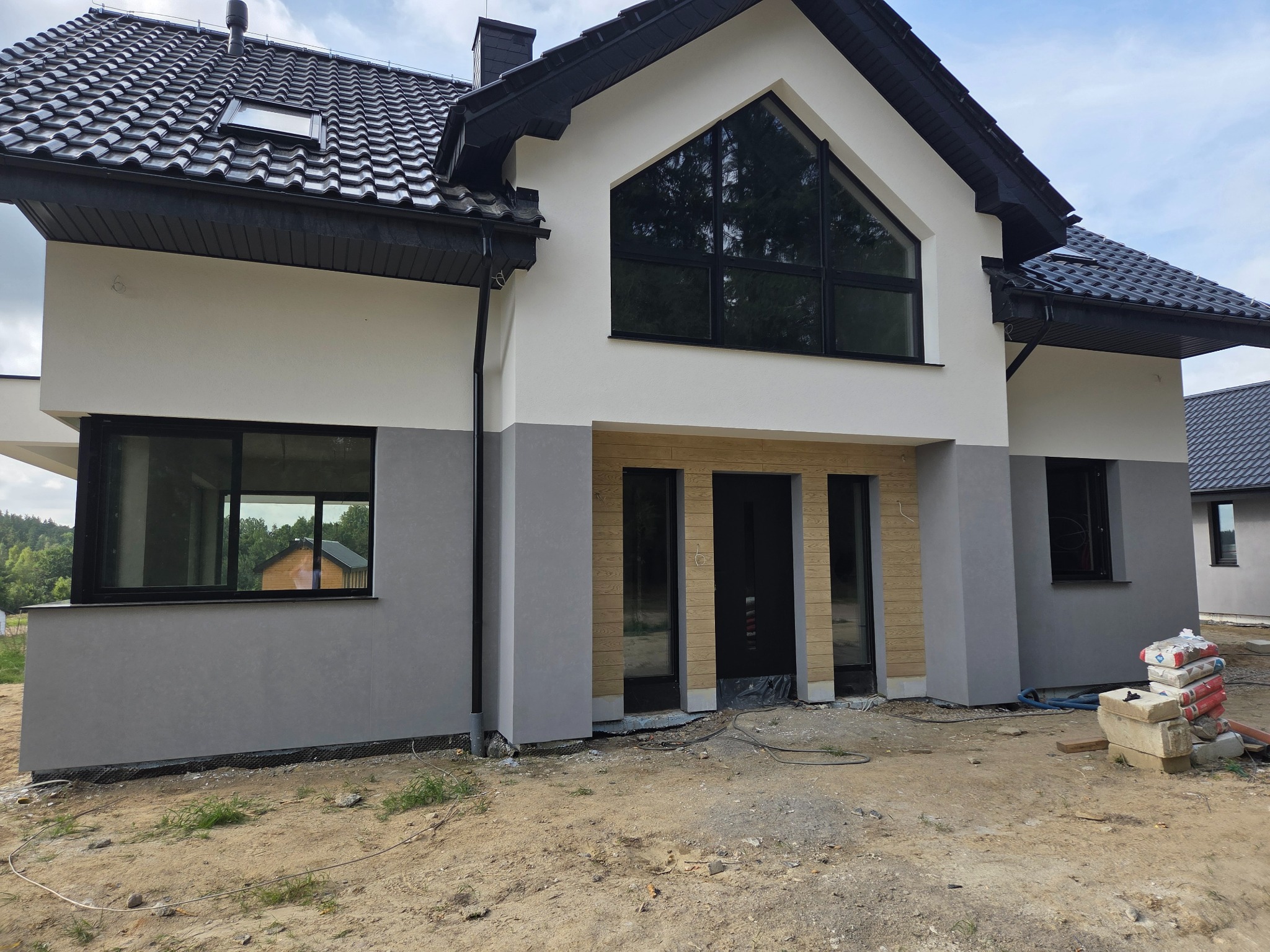 Nowoczesny dom z biało-szarą elewacją i czarną stolarką okienną, ukończony w większości, z widocznymi workami cementu na podwórku.