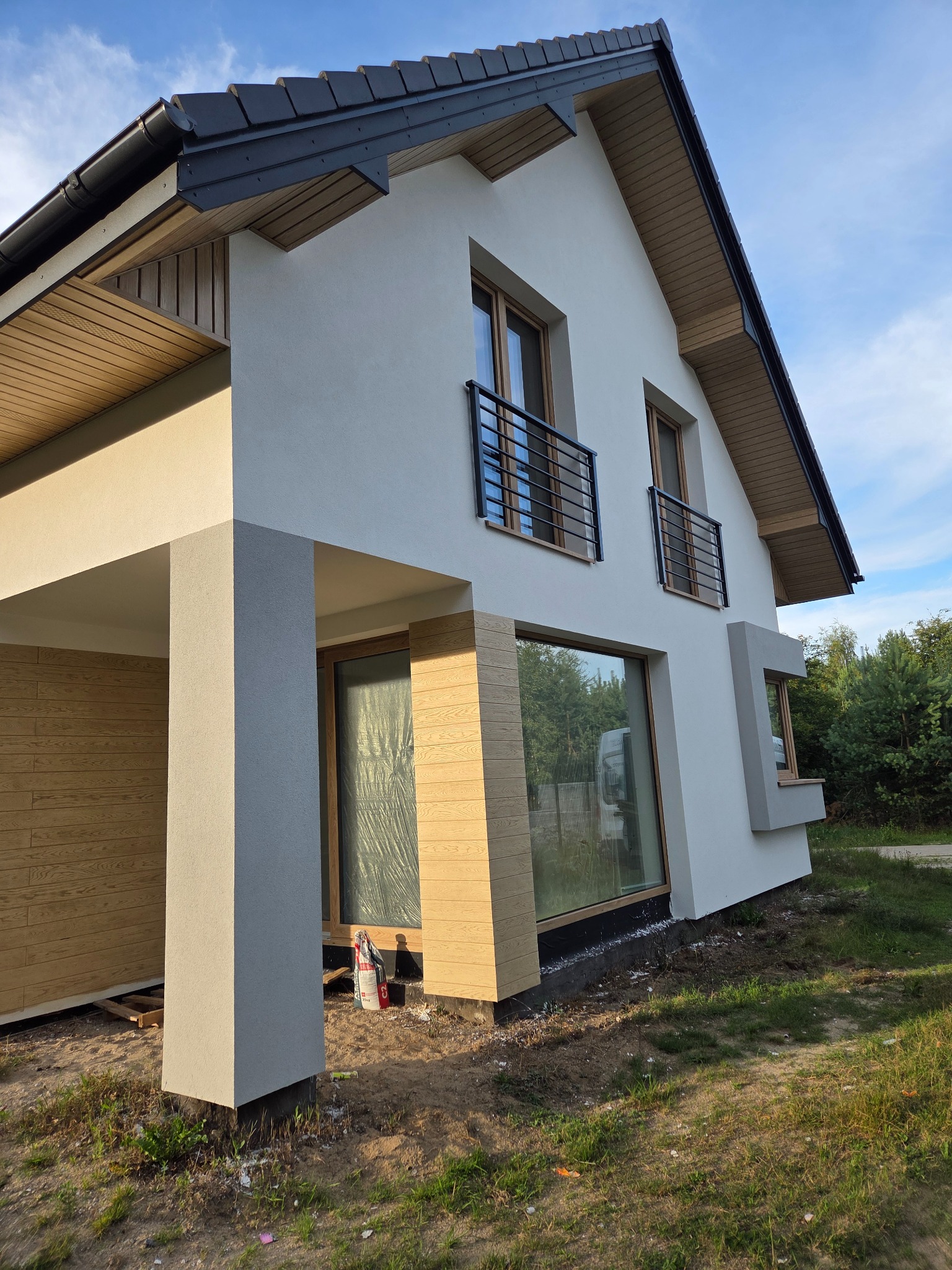 Nowoczesny dom z jasną elewacją, drewnianą okładziną i czarnymi balkonami. Widoczne duże okna i elementy wykończeniowe. Ujęcie z dołu, podkreślające bryłę budynku.