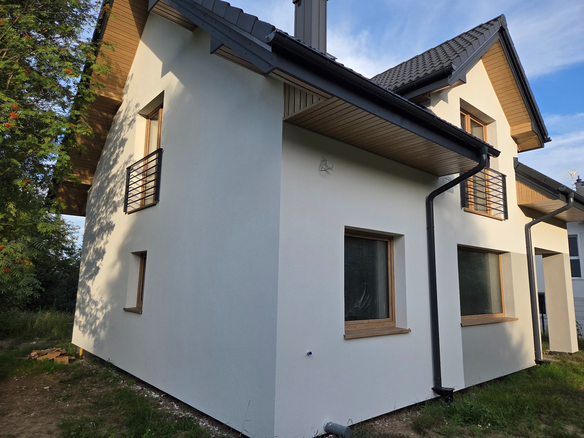 Nowoczesny dom z białą elewacją, drewnianą podbitką i ciemną stolarką okienną. Widoczne elementy drenażu i balkony z czarnymi balustradami. Ujęcie z zewnątrz, w słoneczny dzień.