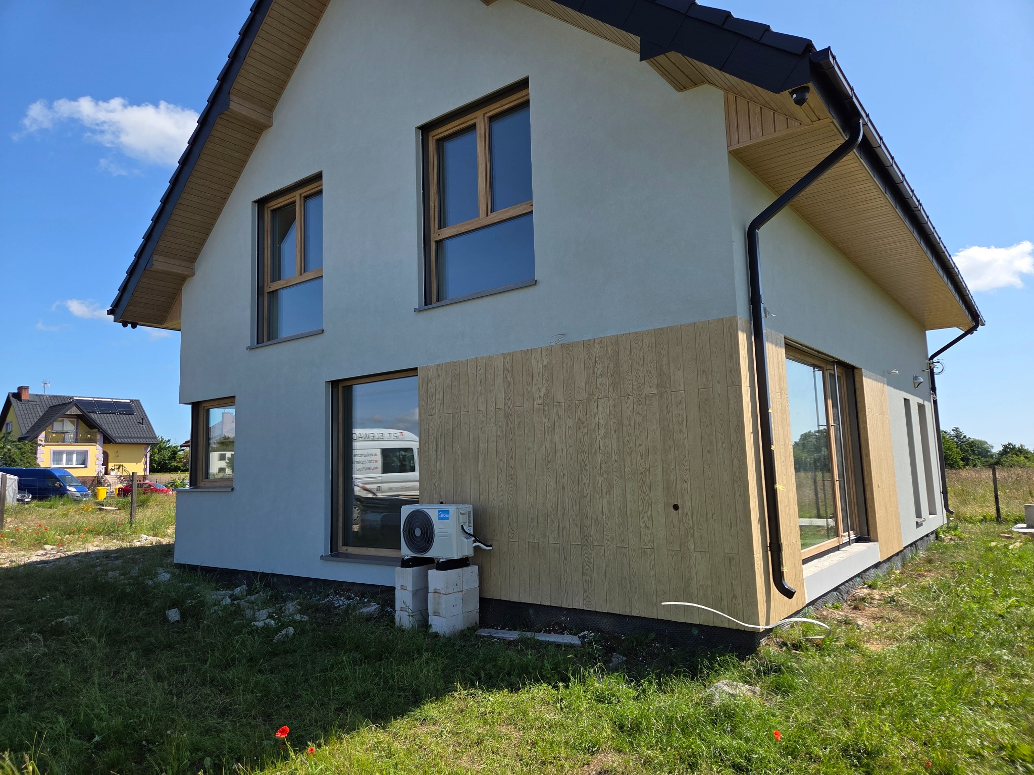 Dwukondygnacyjny dom z oknami w drewnianych ramach i częściowo wykończoną elewacją z drewnianych paneli. Na zewnątrz zamontowany klimatyzator.