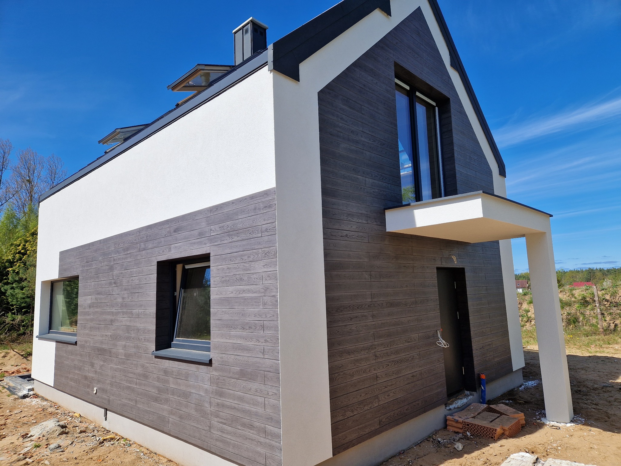 Nowoczesny dom z elewacją z paneli imitujących drewno, białe elementy wykończeniowe, błękitne niebo w tle. Widok z zewnątrz, ukończona realizacja.