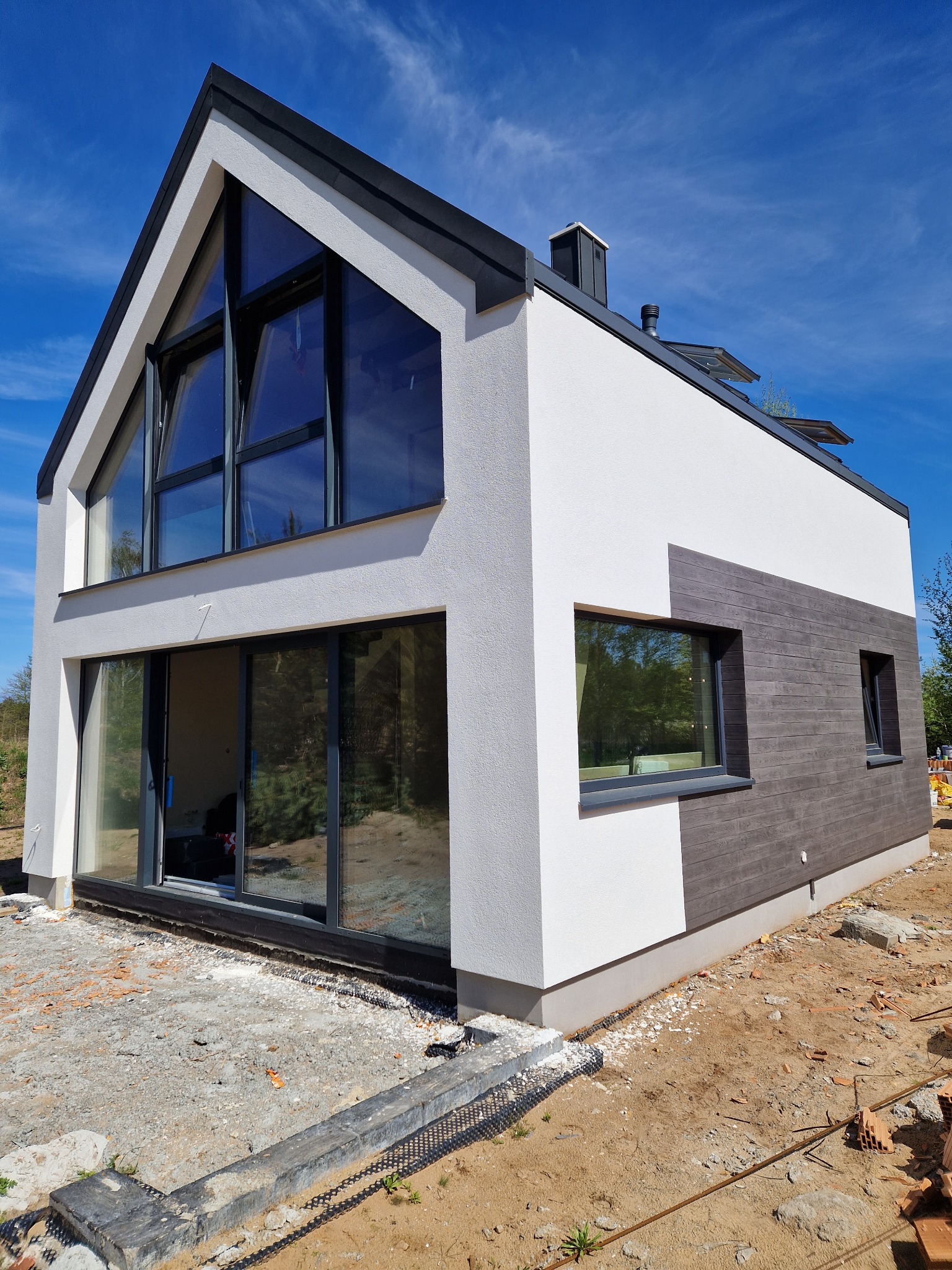 Nowoczesny dom z białą elewacją i elementami drewnopodobnymi, duże okna, niebieskie niebo. Widoczne niedokończone prace ziemne wokół budynku.