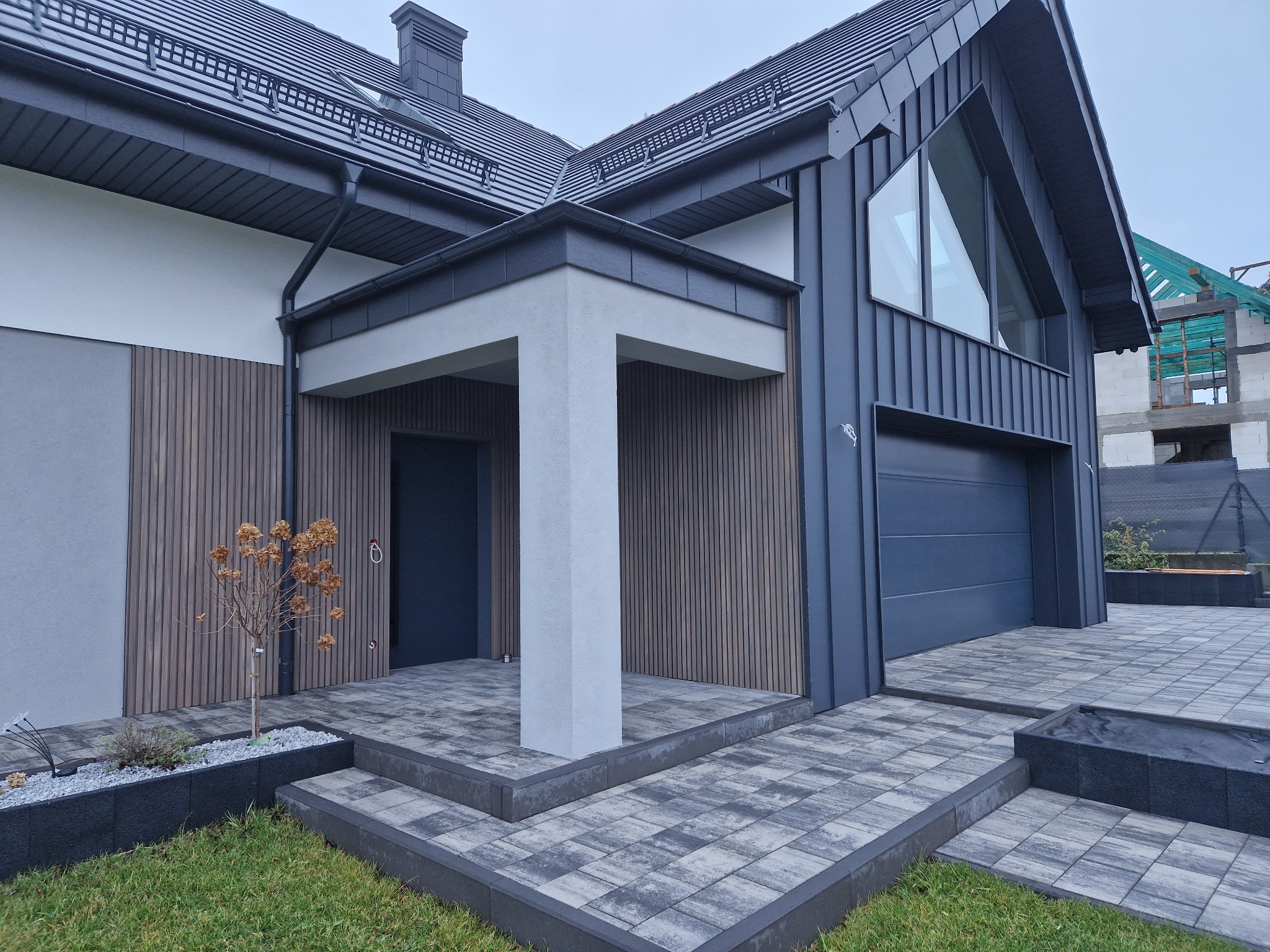 Nowoczesny dom z szarą elewacją, drewnianymi panelami i kostką brukową. Widoczny fragment ogrodu z niską roślinnością i elementami małej architektury.