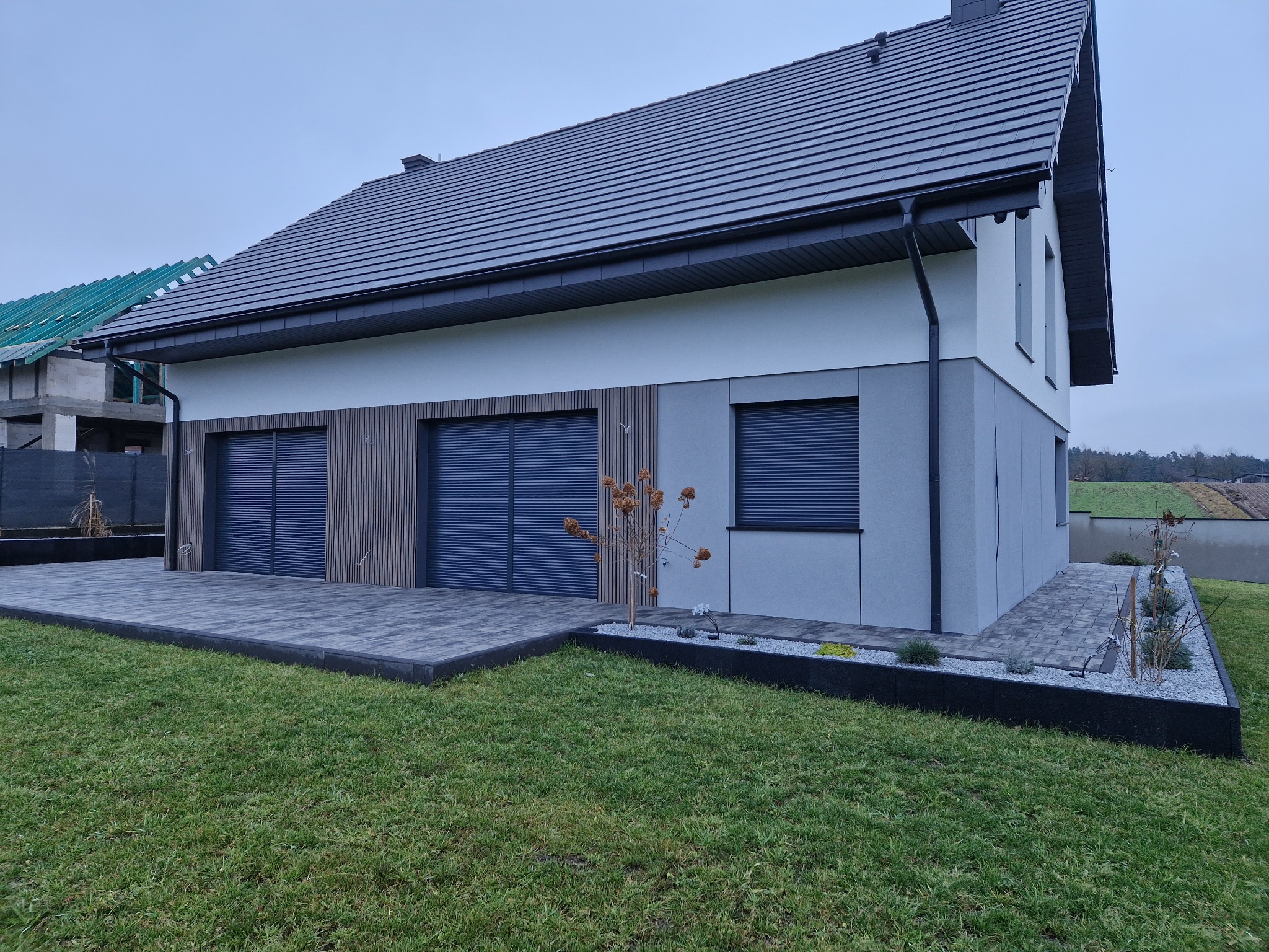 Nowoczesny dom jednorodzinny z szarą elewacją, drewnianymi akcentami i dużym tarasem z kostki brukowej. Widoczny fragment ogrodu i sąsiedni budynek w budowie.