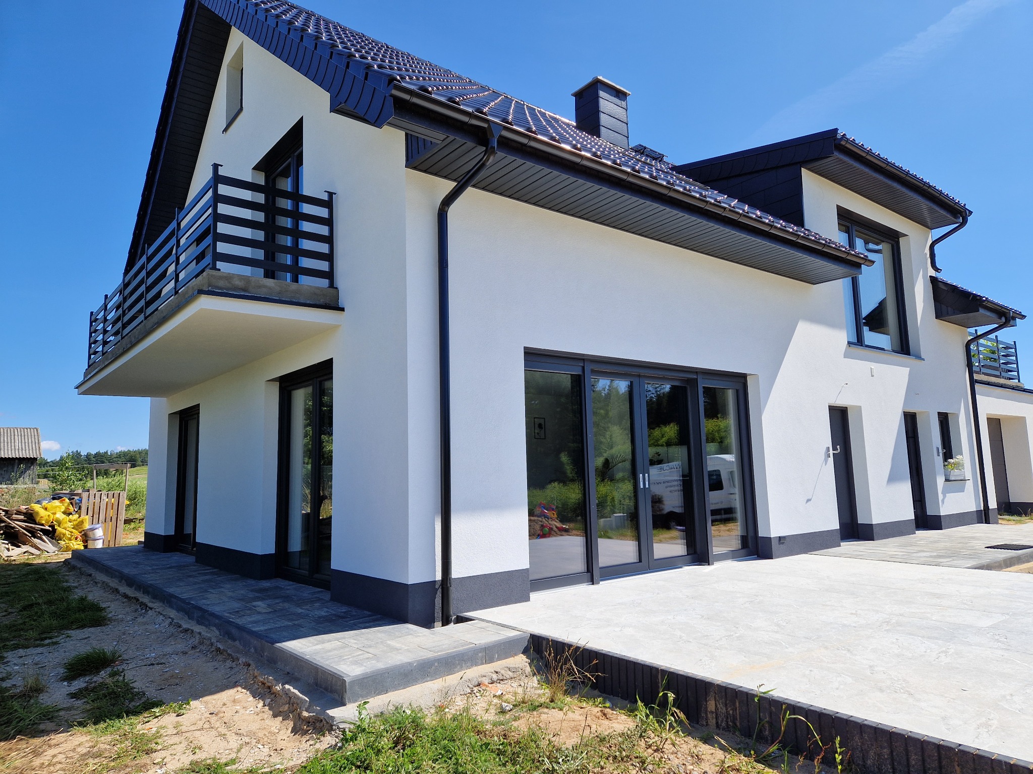 Nowoczesny dom z białą elewacją, czarnym dachem i stolarką. Widoczny balkon z balustradą i taras z szarej kostki. Błękitne niebo w tle.