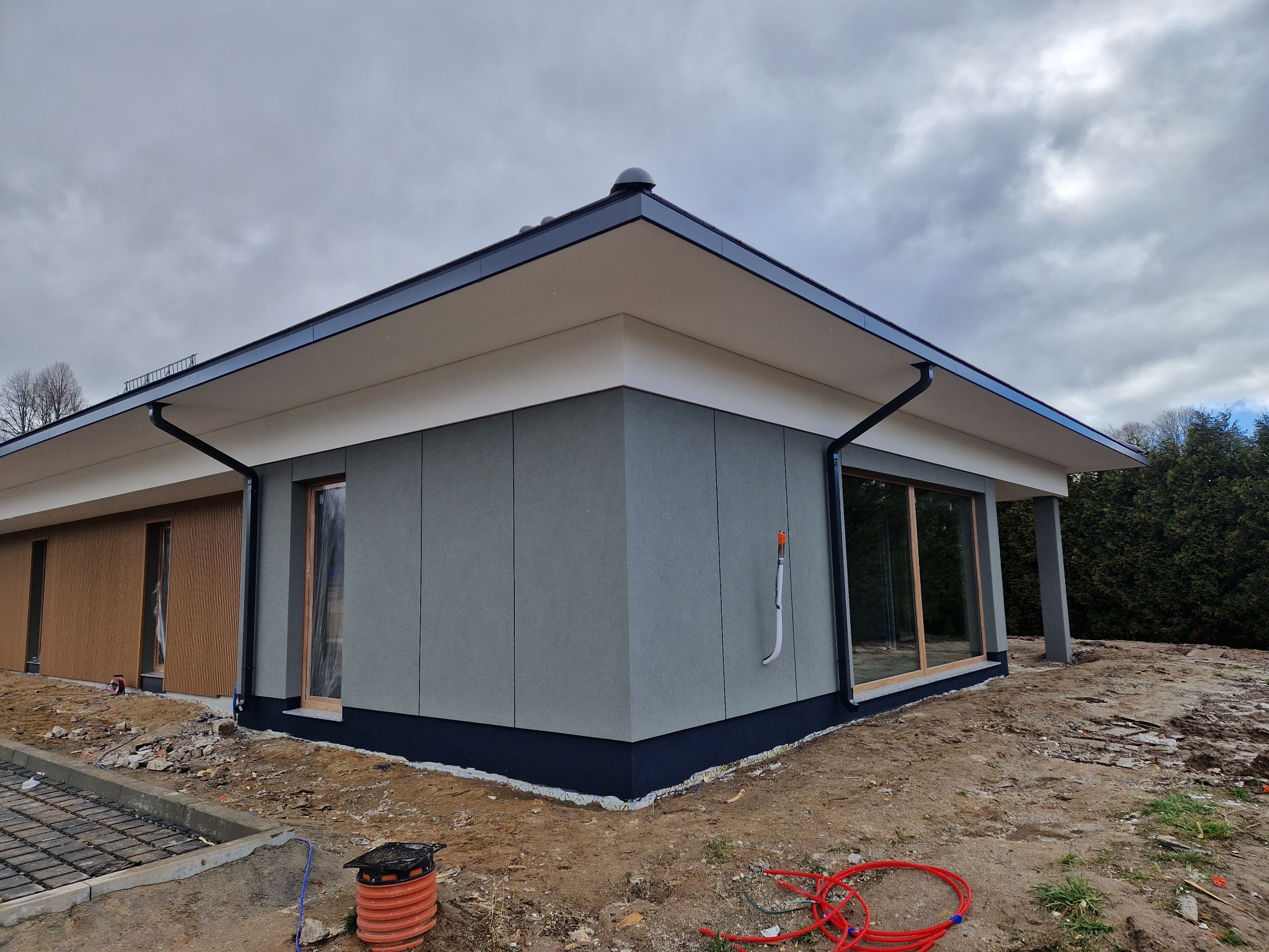 Nowoczesny dom z szarą elewacją panelową, ciemnym dachem i rynnami. Widoczne okna w drewnianych ramach, teren wokół domu w trakcie prac wykończeniowych.