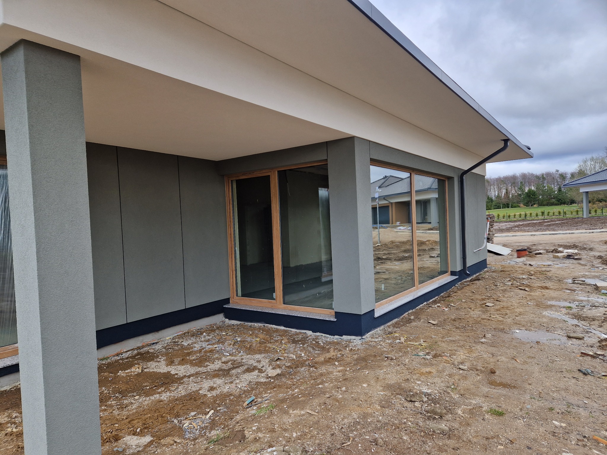 Szara elewacja domu jednorodzinnego z dużymi oknami w drewnianych ramach, widok z zewnątrz, teren wokół domu w trakcie prac wykończeniowych.