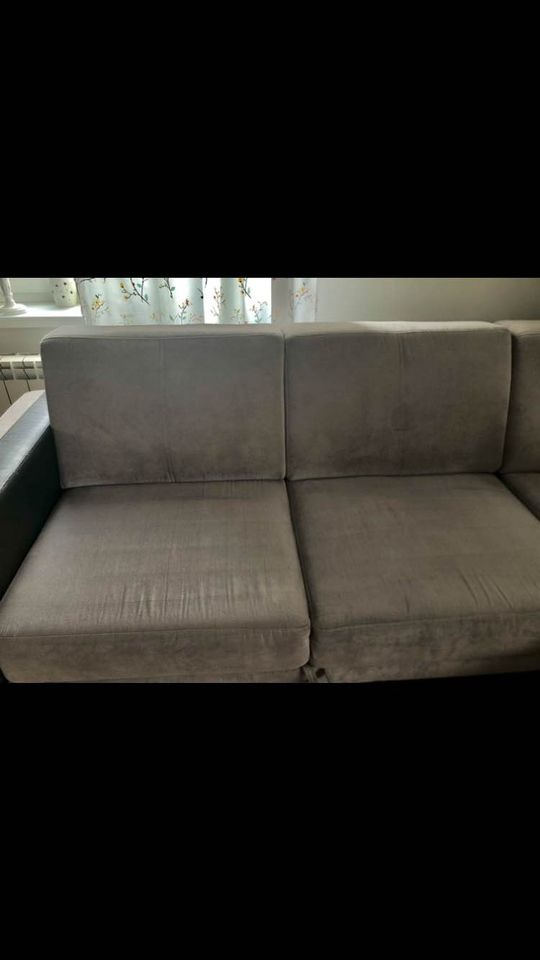 Szara kanapa z widocznymi śladami użytkowania, w tle okno z zasłonami w kwiatowy wzór.