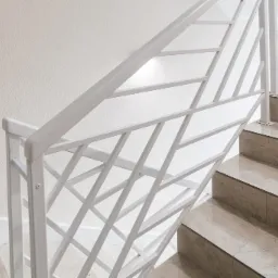 Biała, metalowa balustrada o geometrycznym wzorze na schodach wyłożonych beżowymi płytkami.