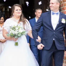 Para młoda wychodzi z kościoła, obsypywana białymi płatkami konfetti, panna młoda w białej sukni z koronkową górą i bukietem, pan młody w granatowym garniturze z muchą, uśmiechnięci.